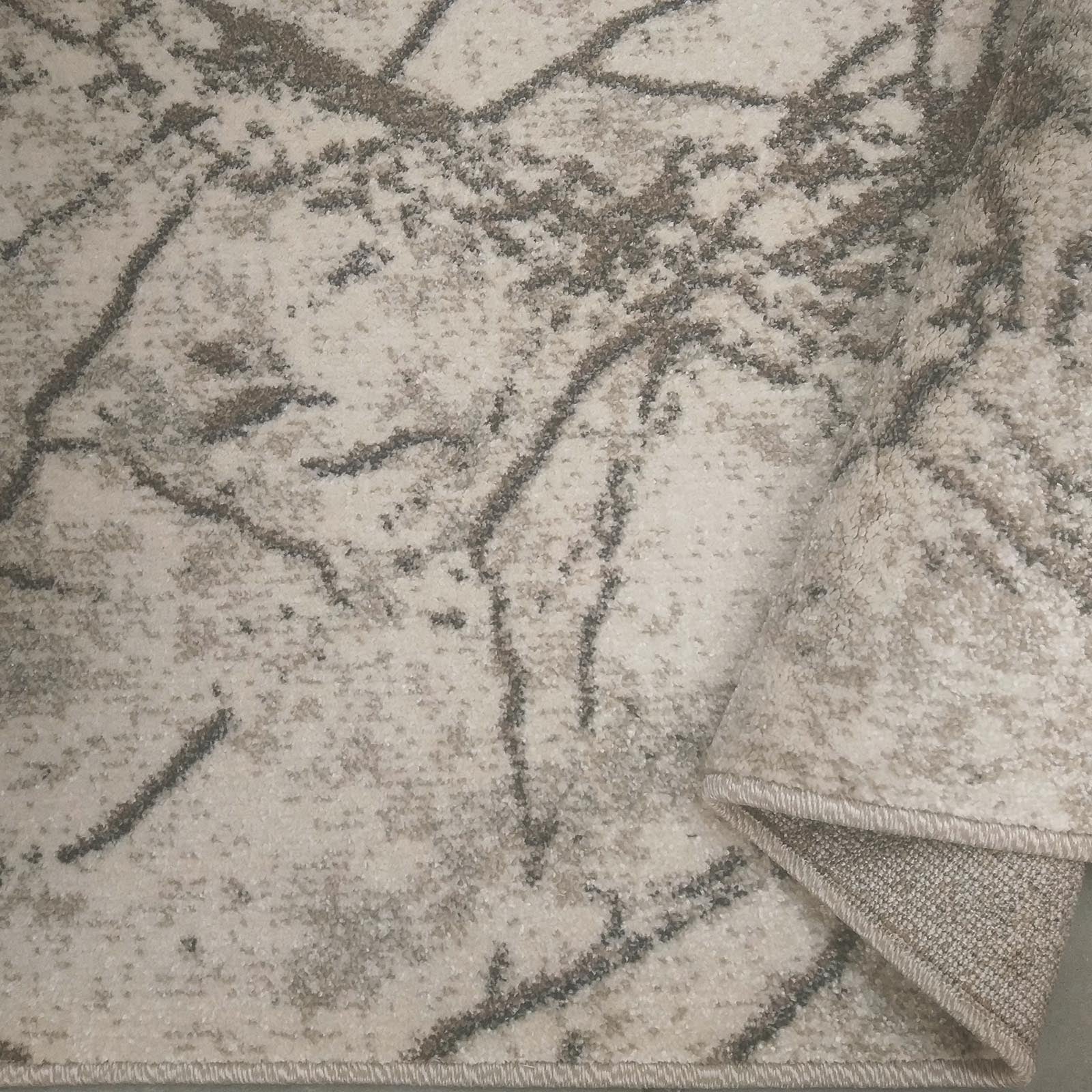 Tapis doux aux motifs abstraits de lignes gris-brun sur un fond crème, avec un revers beige visible sur un coin replié