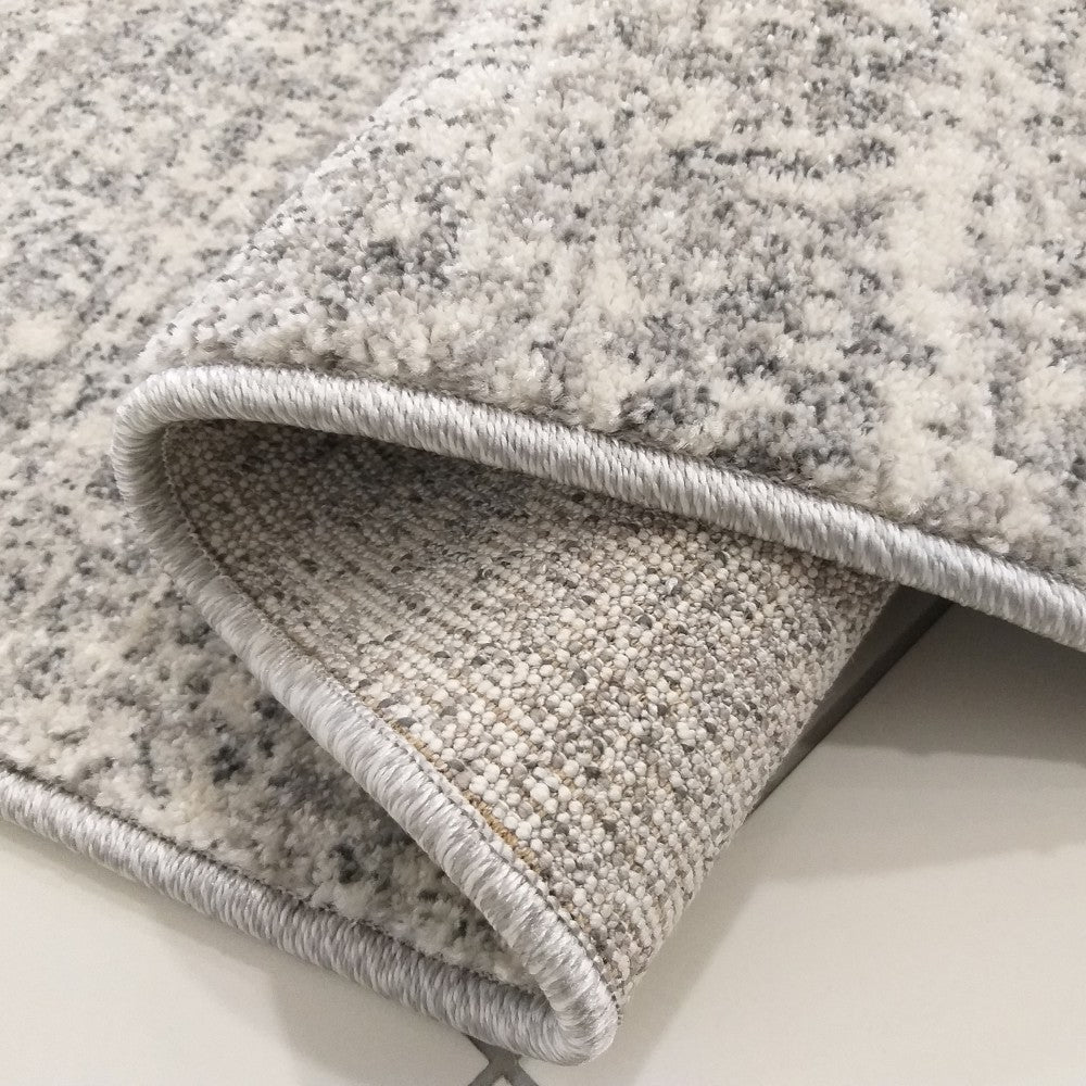 Tapis moelleux aux motifs abstraits gris et blancs, un coin est replié montrant le revers tissé beige et le bord surpiqué gris