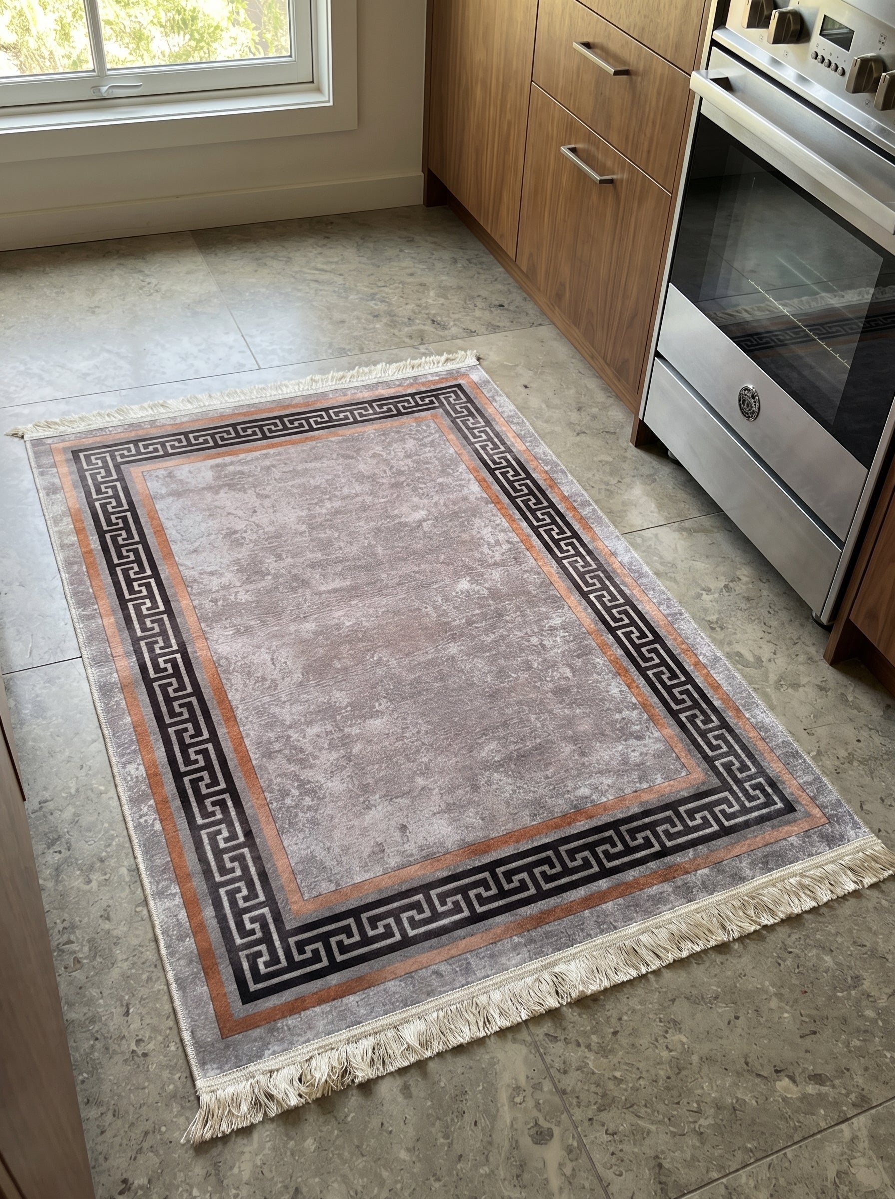 Tapis rectangulaire gris vieilli à motifs méandres noirs et bandes orange, avec franges claires, posé sur un sol carrelé dans une cuisine