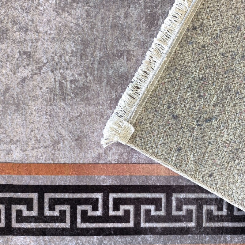 Tapis gris moucheté, avec bordure à motif méandre noir et gris, et bandes marron et grises. Un coin du tapis est relevé, montrant son envers tissé quadrillé vert clair et des franges claires