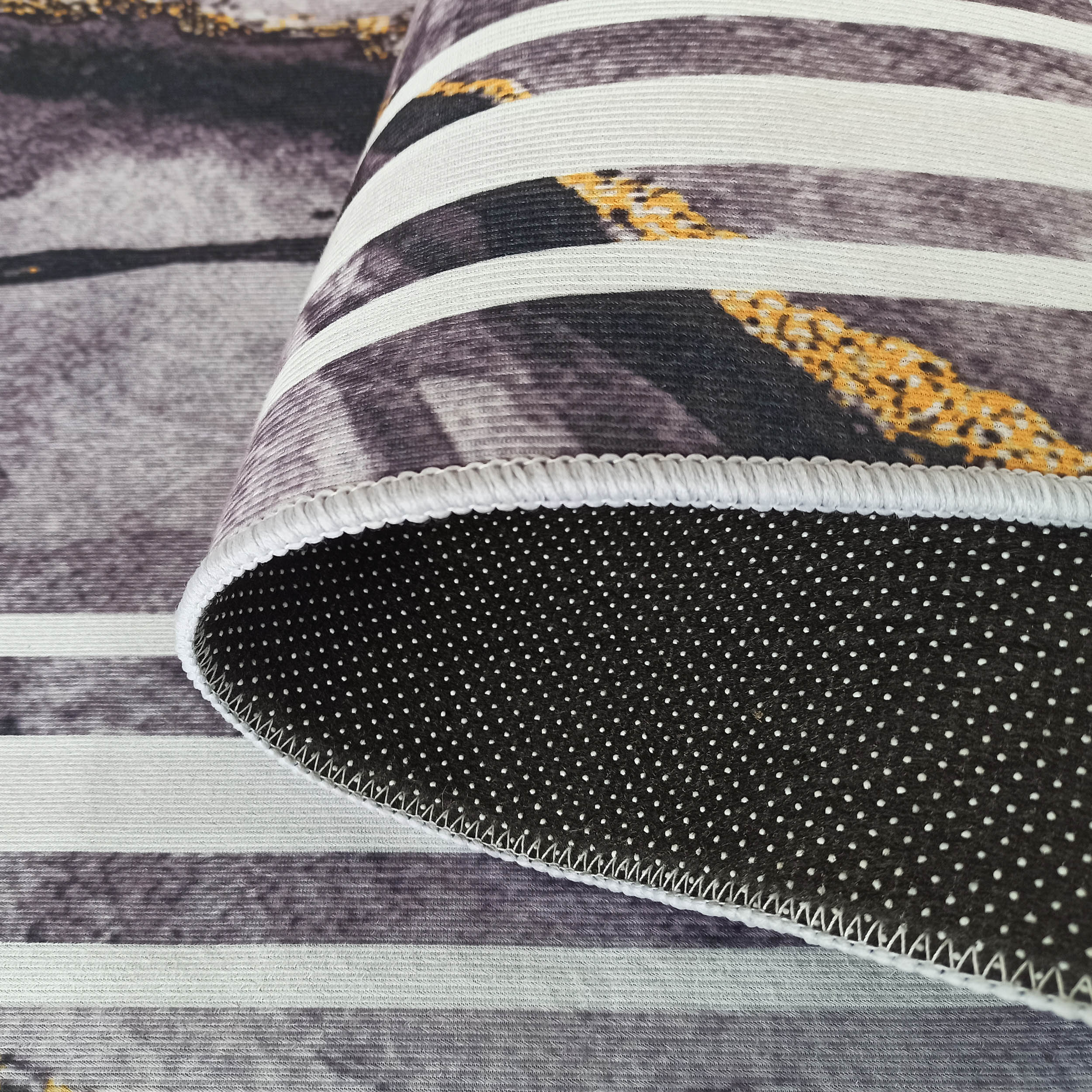 Tapis doux à rayures horizontales grises, blanches et tachetées de jaune. L'envers est noir à pois antidérapants blancs