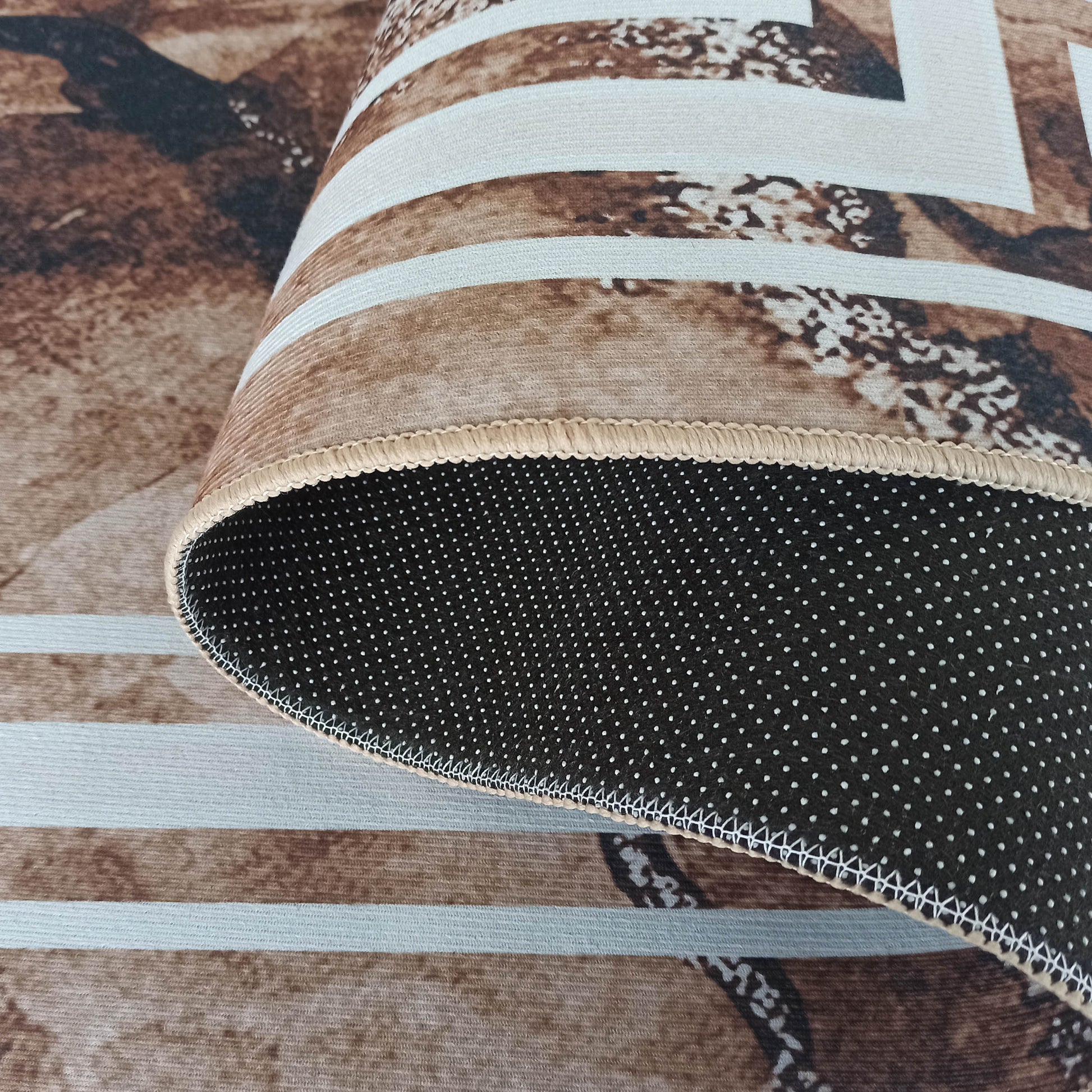 Un tapis roulé montrant son côté supérieur avec motifs abstraits marron et beige, et son envers noir à pois blancs avec une bordure beige