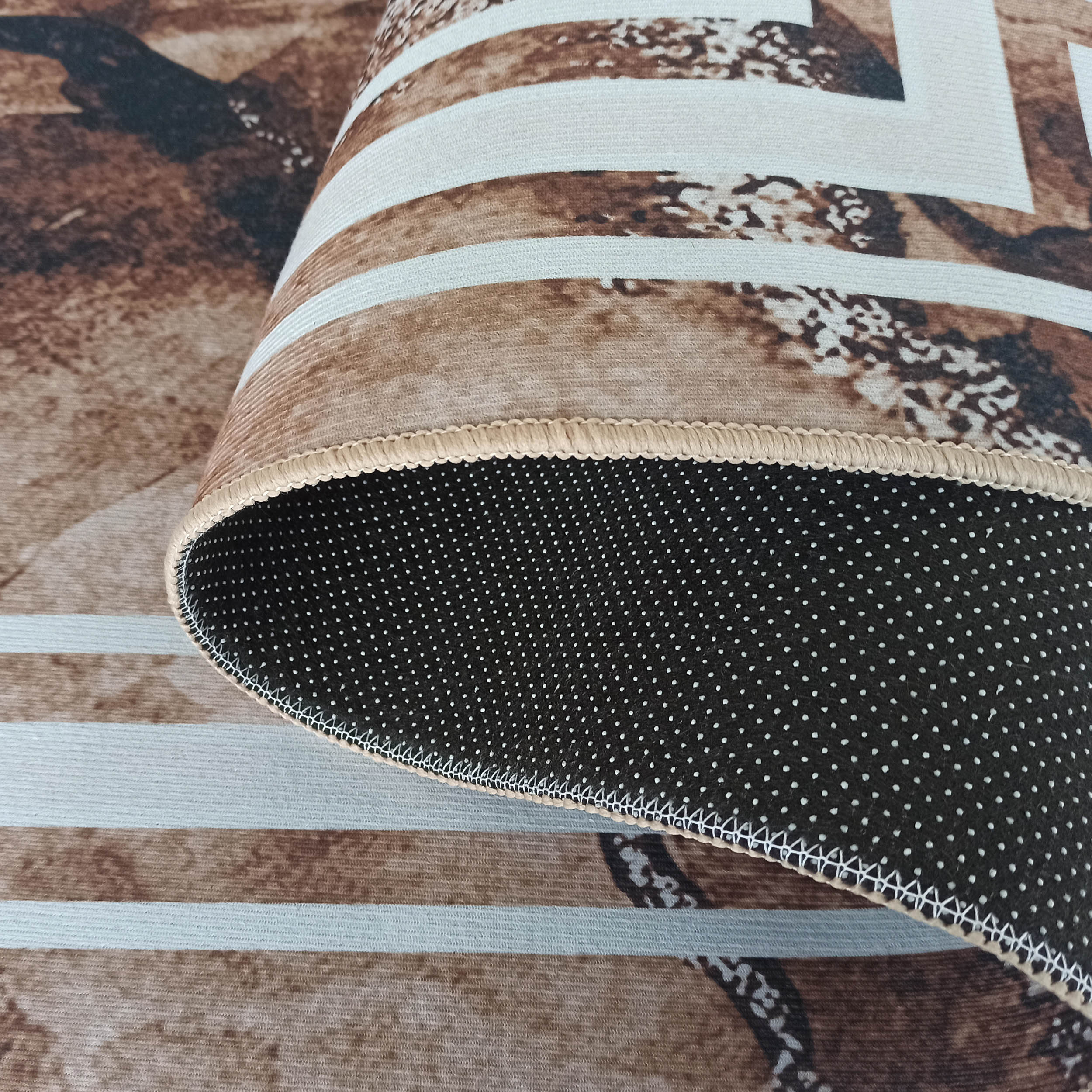 Un tapis roulé montrant son côté supérieur avec motifs abstraits marron et beige, et son envers noir à pois blancs avec une bordure beige