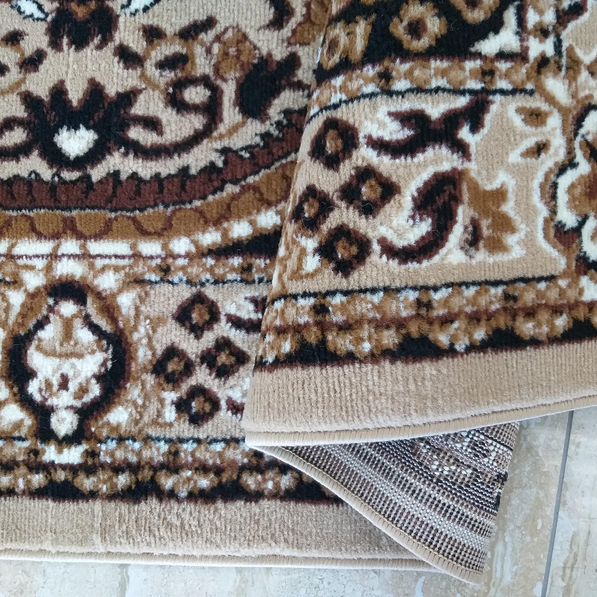 Tapis à poils courts beige avec des motifs ornementaux marron, noirs et blancs, partiellement roulé sur un carrelage