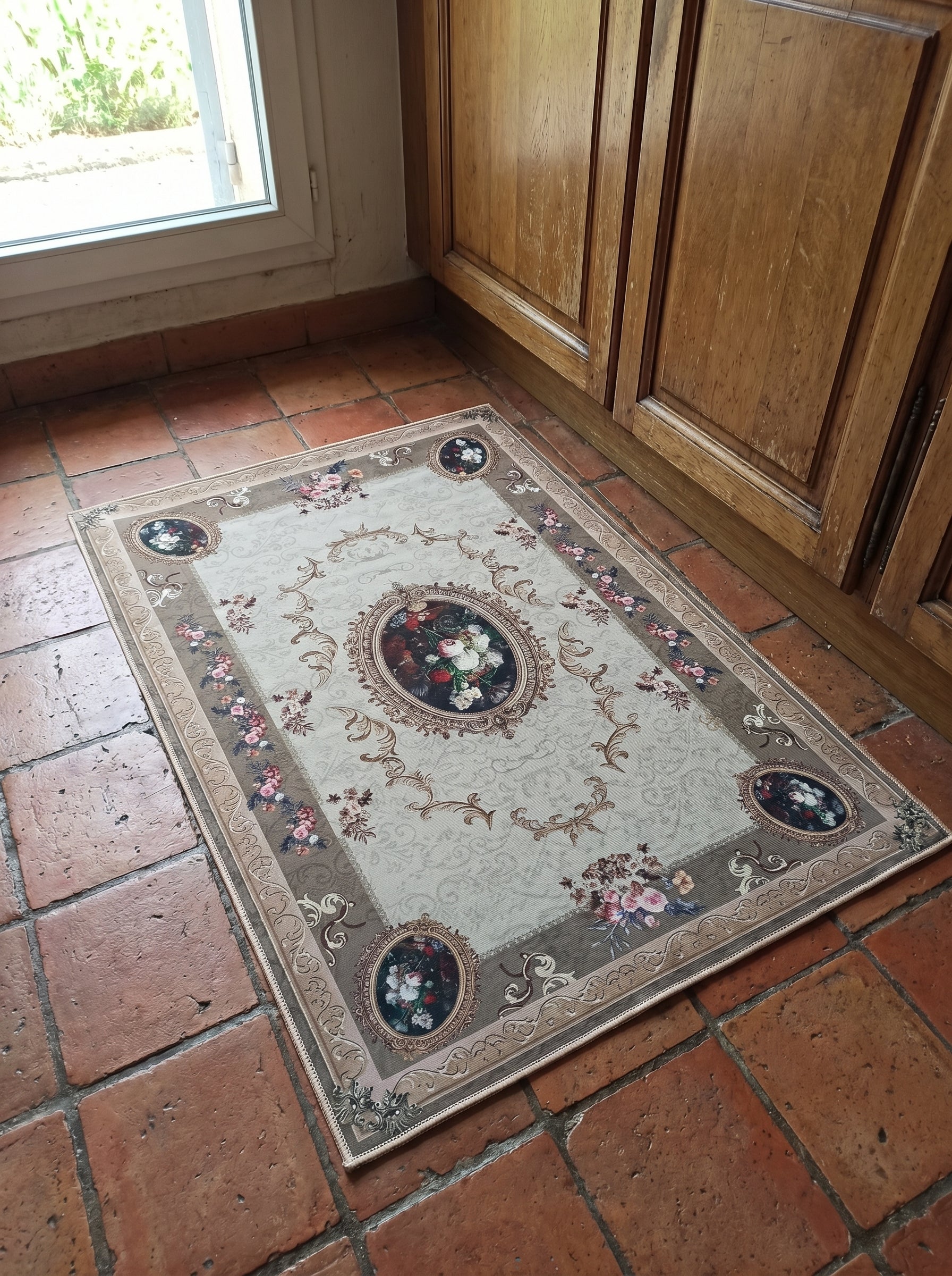 Tapis rectangulaire beige à motifs arabesques dorés, avec médaillons et bordures florales colorées, sur des tomettes près d'un meuble en bois