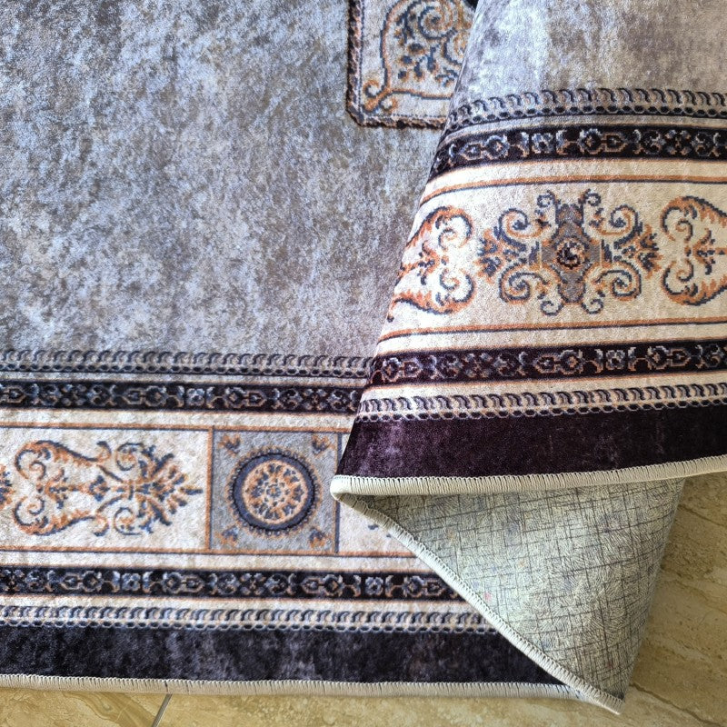 Tapis gris-beige chiné avec une bordure classique aux motifs bleus, bruns et crème, bordée de marron foncé. Le tapis est partiellement roulé