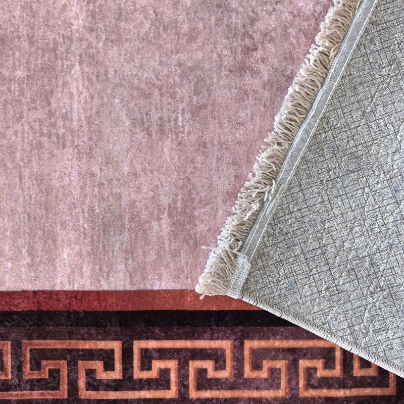 Tapis rose poudré à texture veloutée avec bordure marron et méandres ocre. Un coin replié dévoile l'envers gris texturé et les franges claires