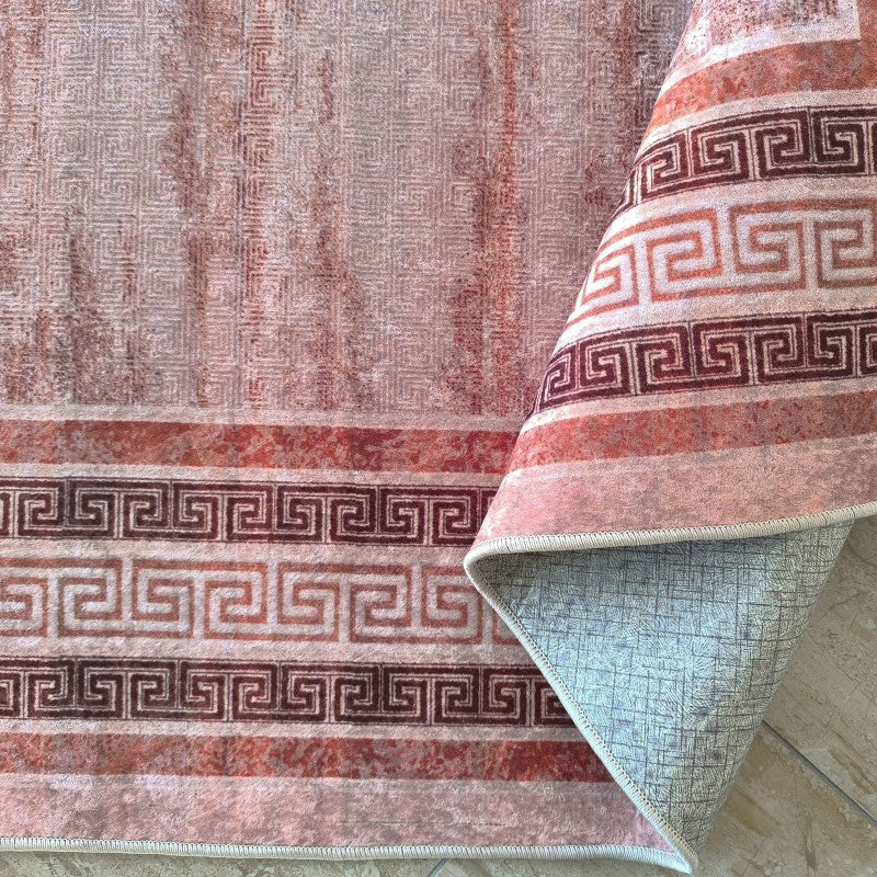 Tapis texturé rose orangé avec motifs géométriques subtils et bandes à méandre grec marron, un coin est replié