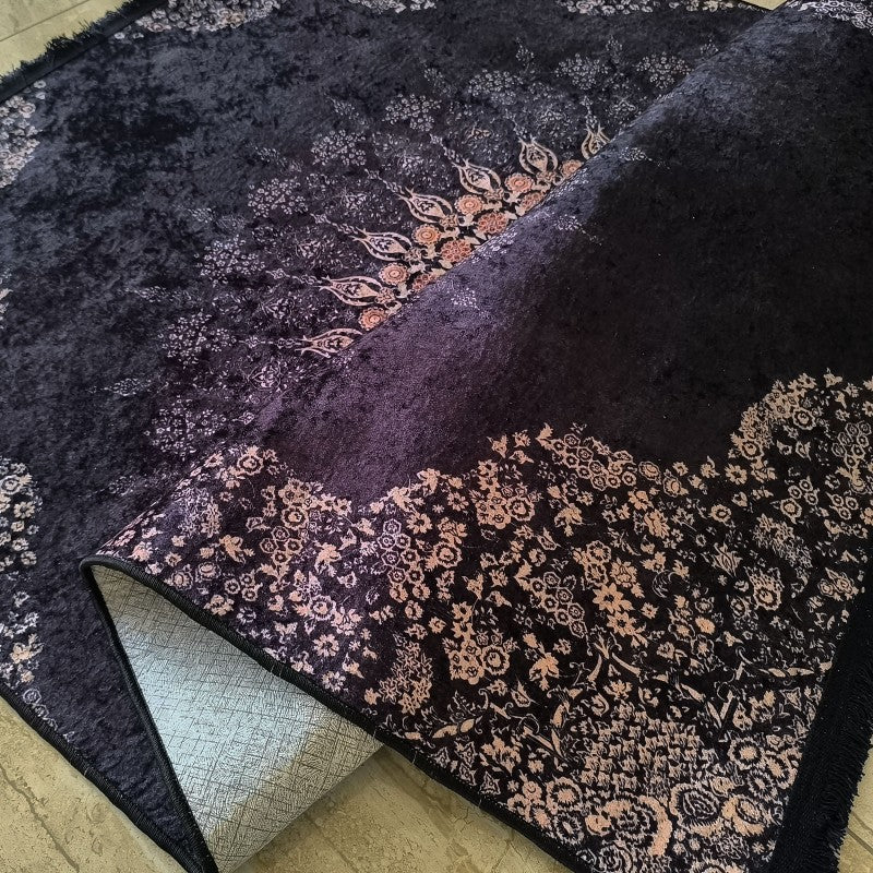 Tapis velouté violet foncé aux motifs floraux et arabesques beige-rosé, avec franges, un coin plié montre son envers quadrillé