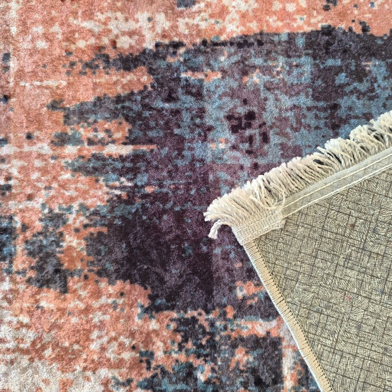 Tapis doux aux motifs abstraits mêlant des tons de pêche, bleu sarcelle et violet. Une frange crème et le dos quadrillé gris du tapis sont visibles