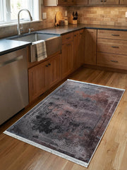 Tapis rectangulaire aux teintes anthracite, rouille et noir, avec motif abstrait et bordure ornée de franges, sur le plancher d'une cuisine