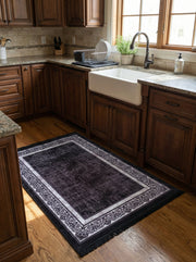 Tapis rectangulaire noir avec un motif central usé et une bordure claire ornée de volutes sombres sur parquet de cuisine