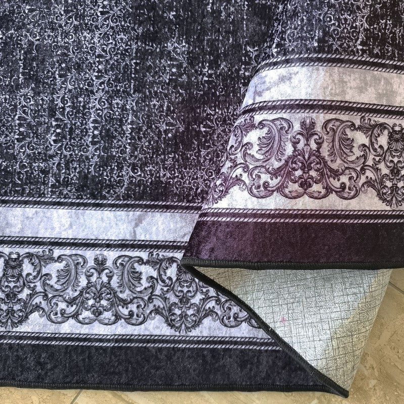Tapis velouté gris foncé aux motifs baroques argentés et pourpres, partiellement roulé pour révéler son envers texturé
