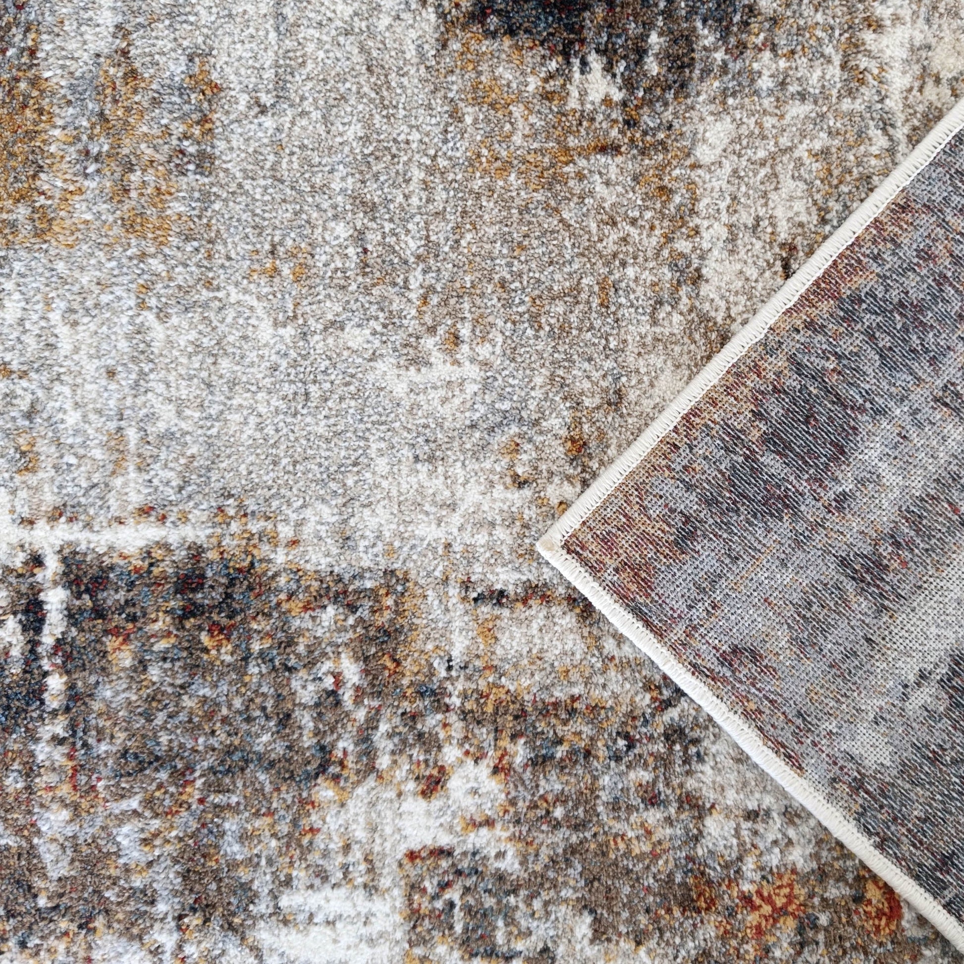 Surface de tapis doux aux motifs abstraits gris, blanc, marron, ocre et rouge, avec un coin relevé sur son dos tissé