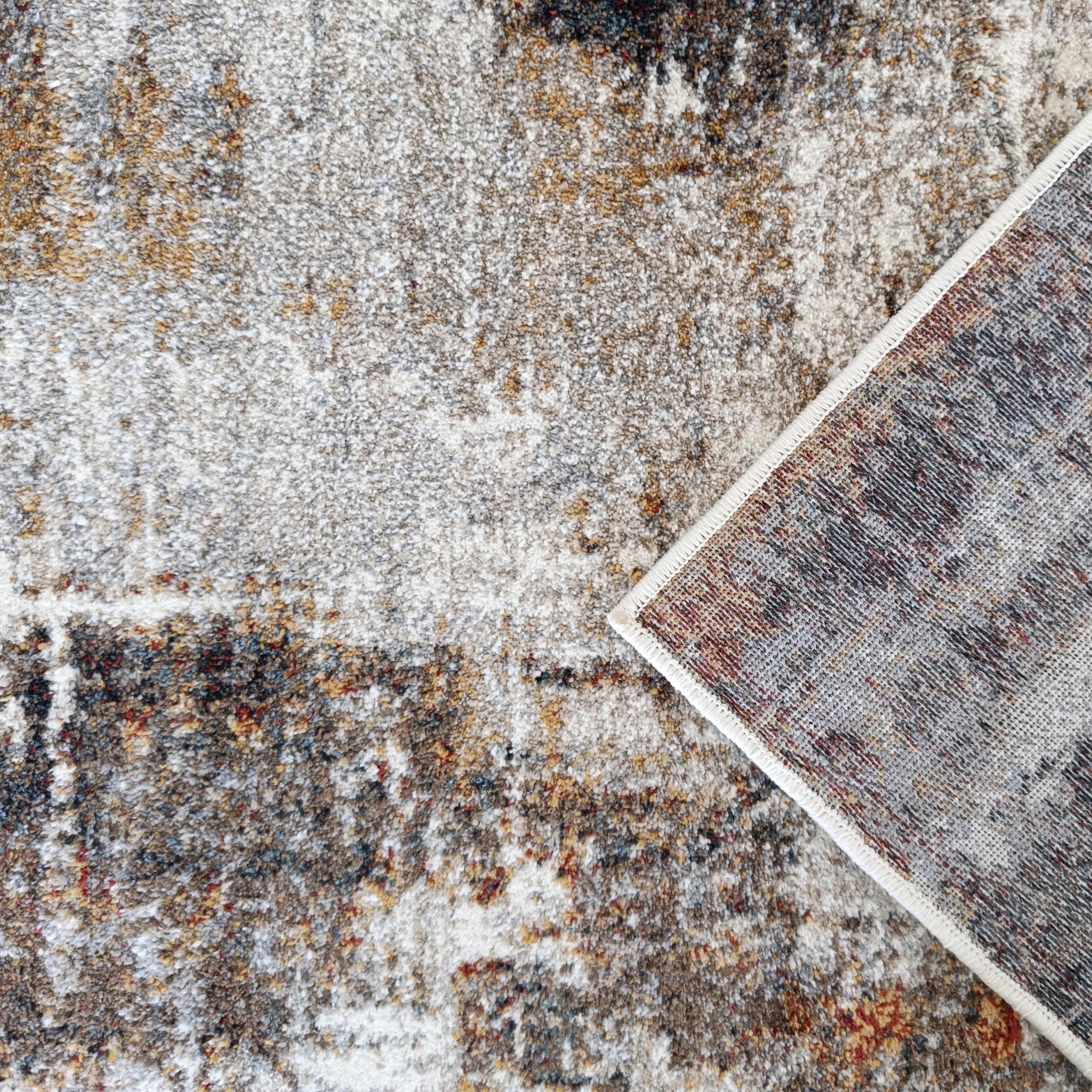 Surface de tapis doux aux motifs abstraits gris, blanc, marron, ocre et rouge, avec un coin relevé sur son dos tissé