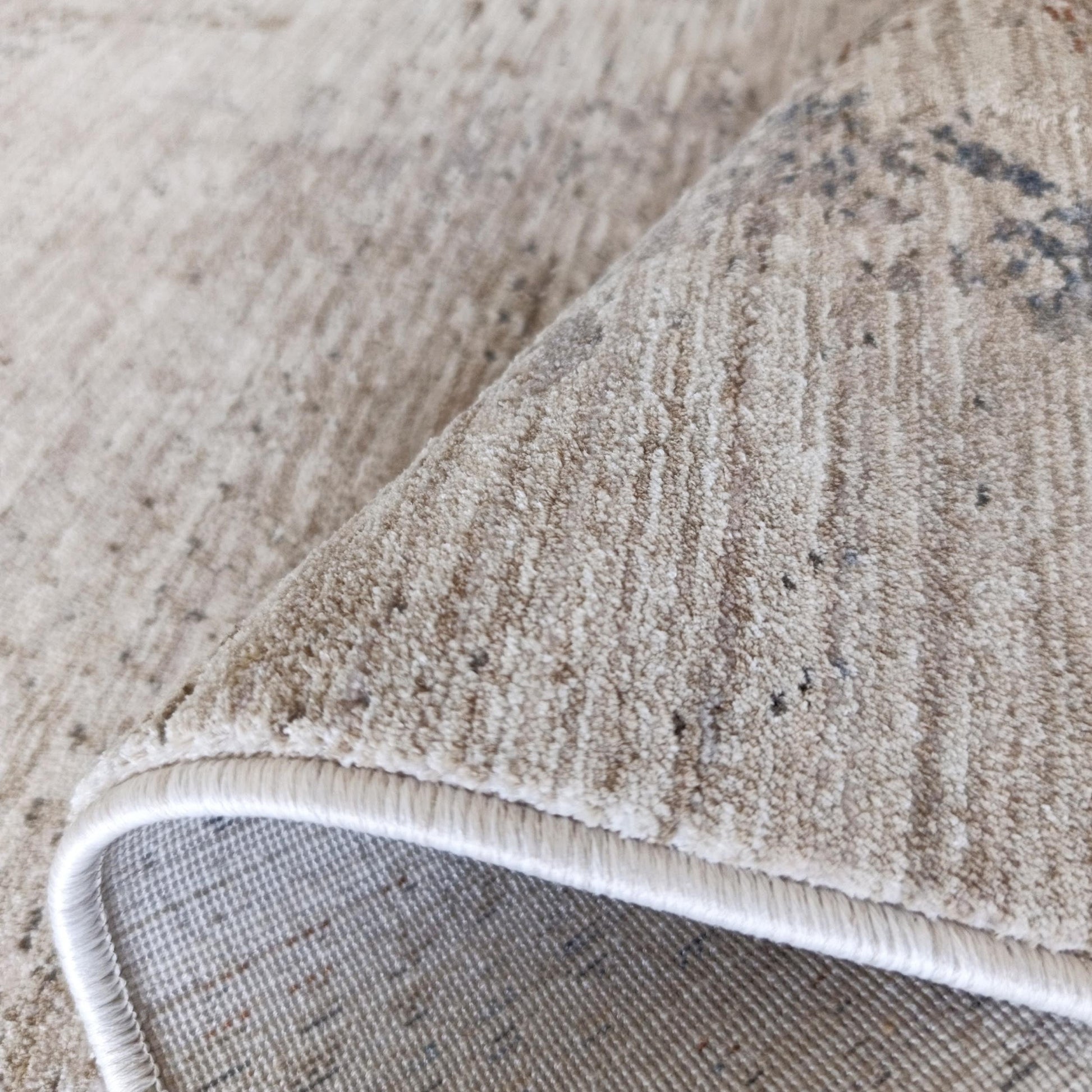 Tapis doux beige clair à motifs abstraits gris foncé, partiellement roulé pour montrer sa bordure blanche et son envers tissé