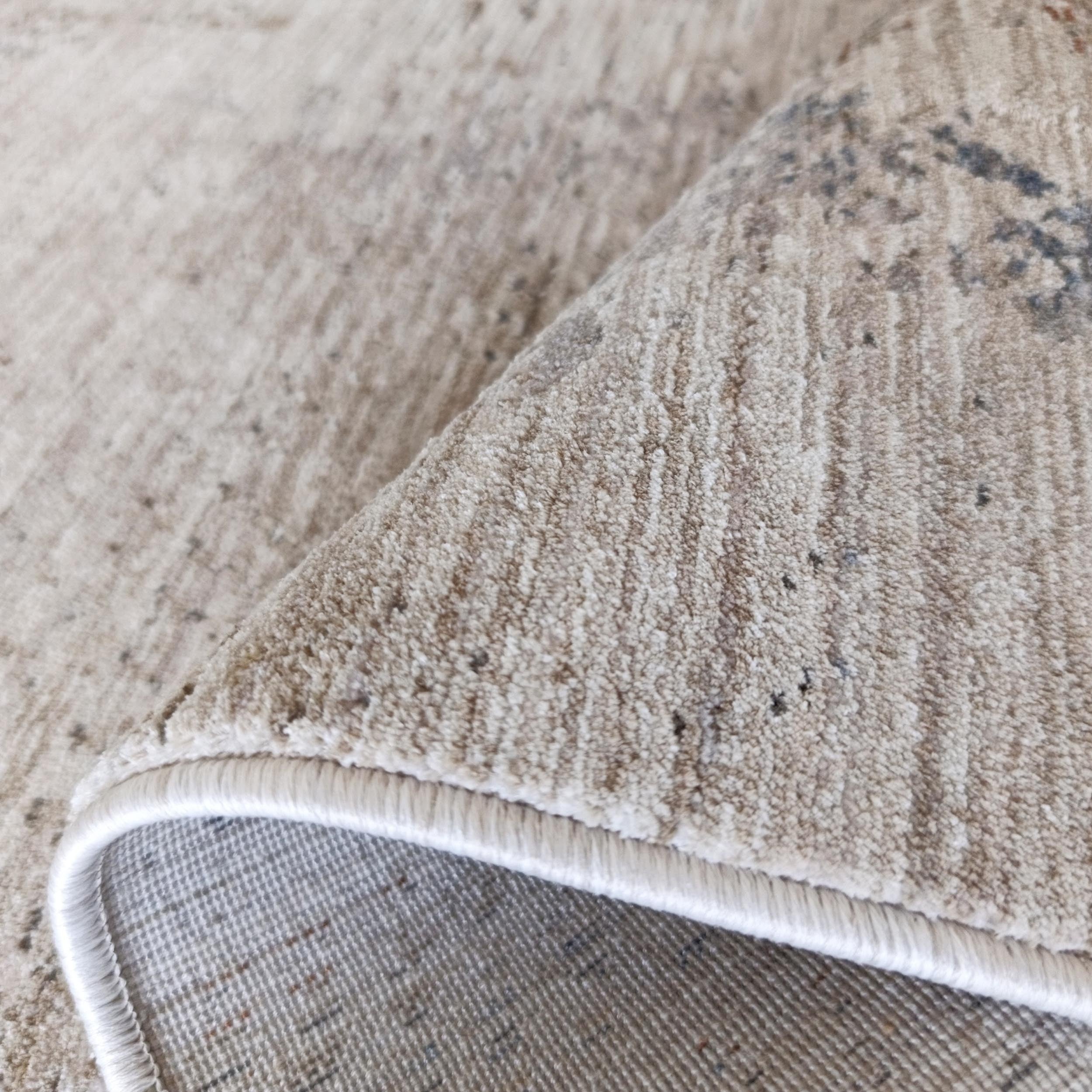 Tapis doux beige clair à motifs abstraits gris foncé, partiellement roulé pour montrer sa bordure blanche et son envers tissé