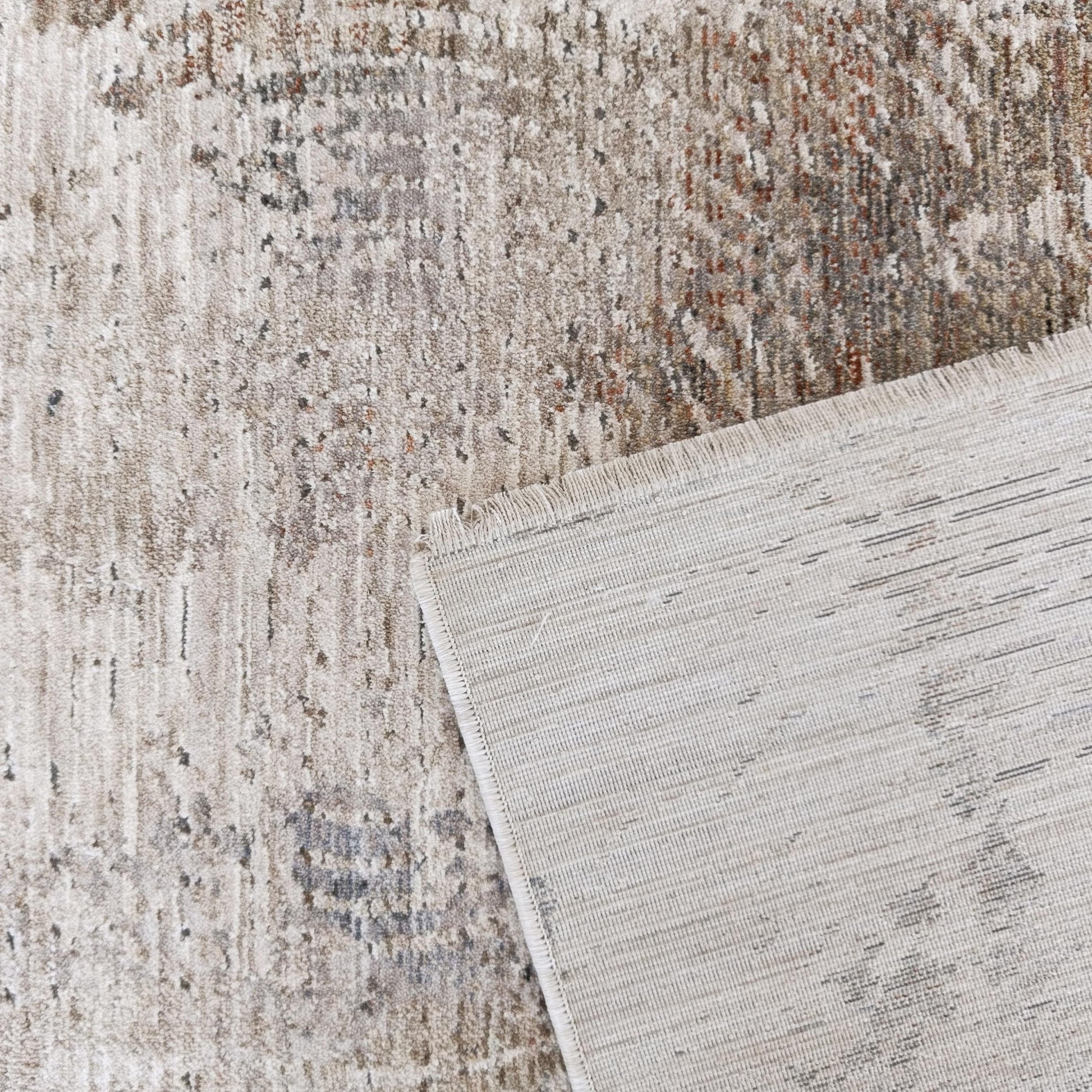 Un tapis texturé abstrait beige, gris, rouille, un coin relevé montre son envers tissé clair et franges