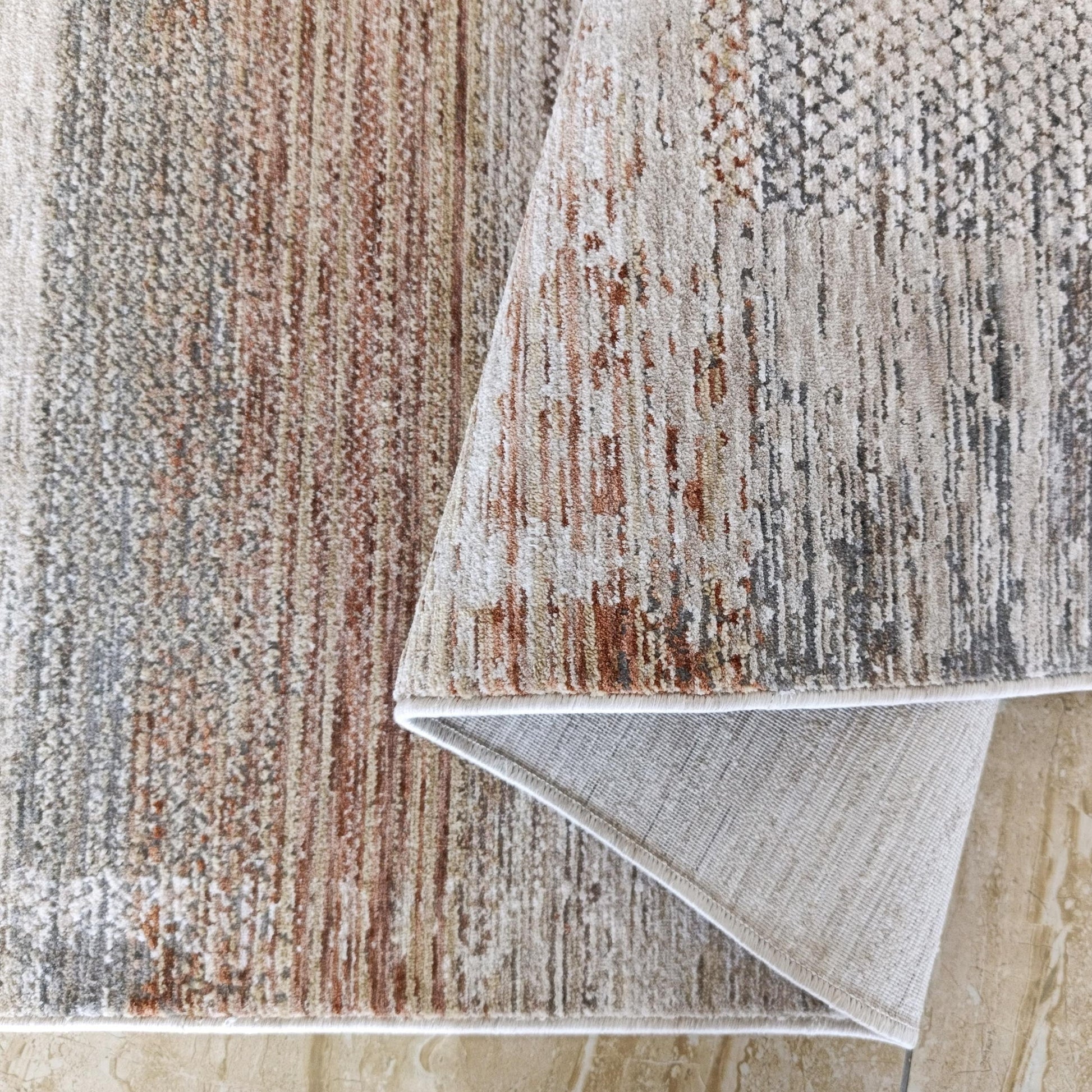 Tapis aux motifs abstraits beiges, gris et rouille, replié sur un sol en marbre clair. Son envers uni est visible
