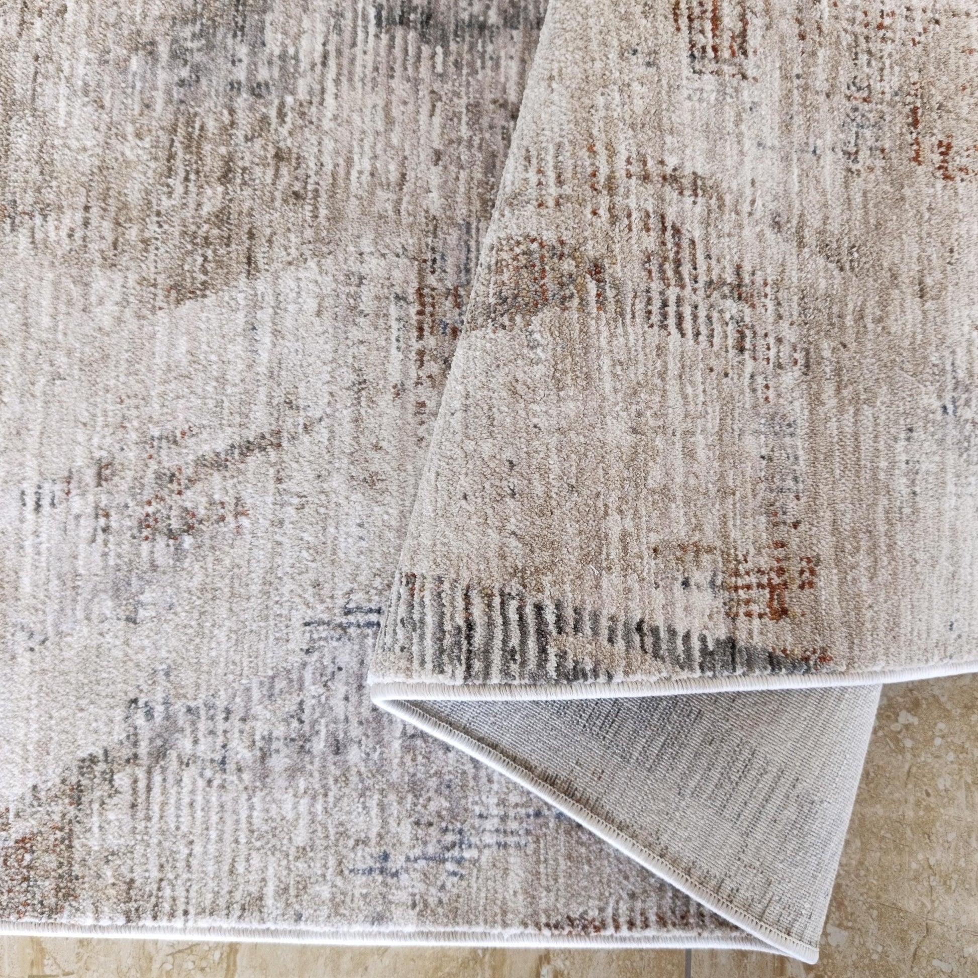 Tapis à motifs abstraits beiges, gris et bruns-rouille, replié sur un carrelage clair marbré, montrant son revers tissé