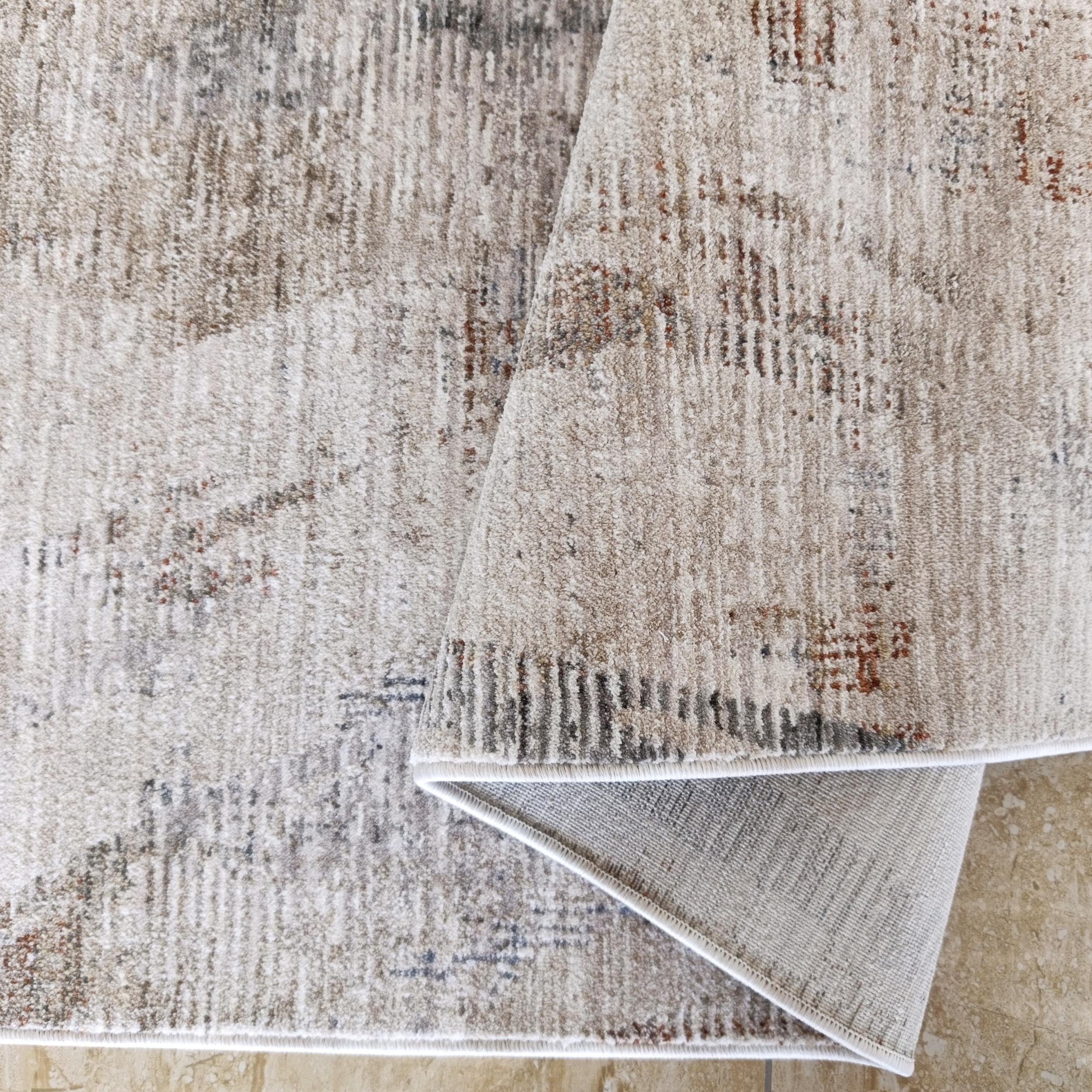 Tapis à motifs abstraits beiges, gris et bruns-rouille, replié sur un carrelage clair marbré, montrant son revers tissé