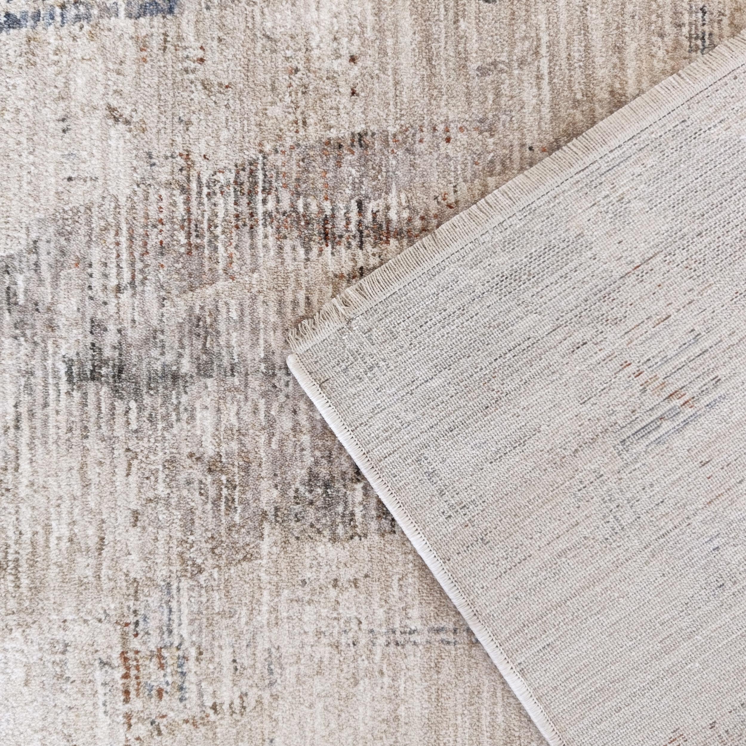 Tapis doux beige clair et gris à motif abstrait, partiellement plié révélant son envers tissé et une frange claire