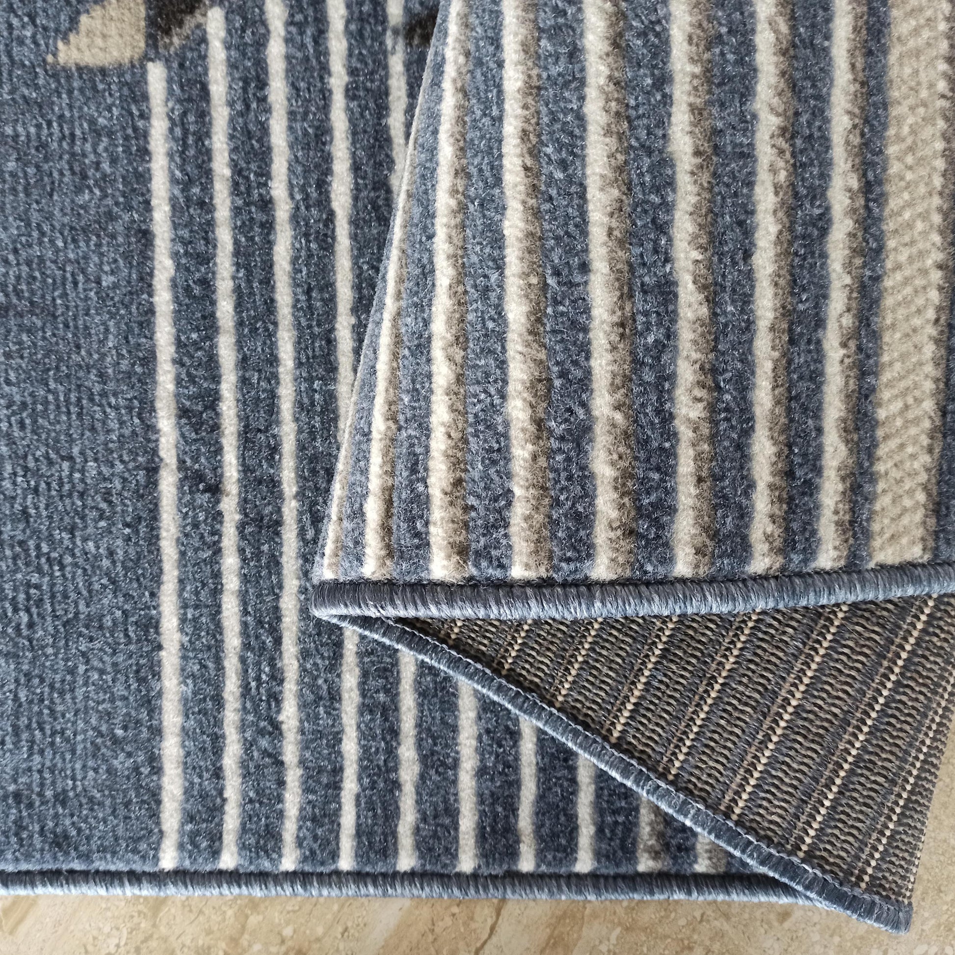 Tapis à poil ras avec des rayures verticales bleu-gris et beige clair. Un coin est relevé, exposant le dos tissé et un liseré