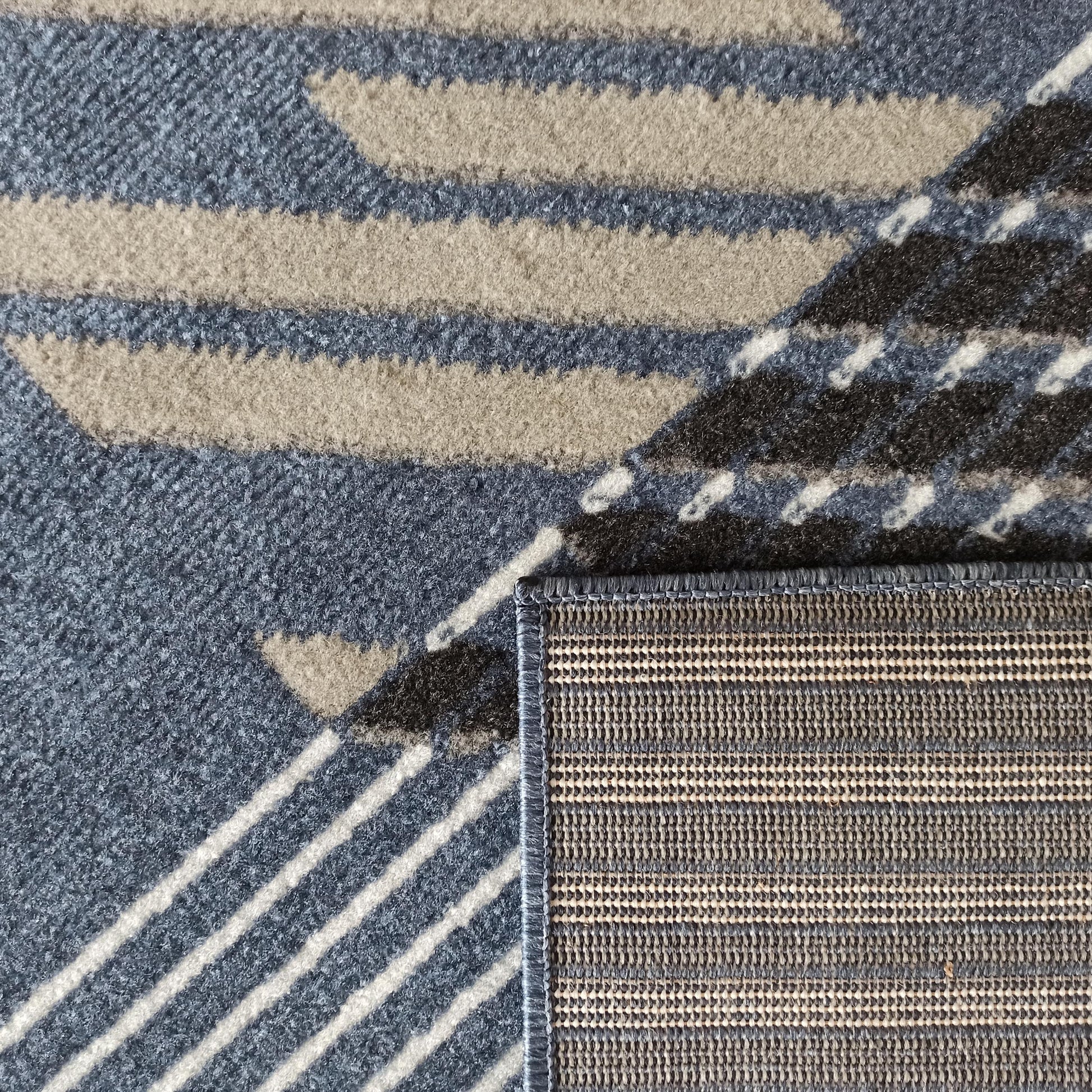 Tapis en tissu à motifs géométriques bleus, beiges, noirs et blancs. Un coin relevé montre l'envers tissé rayé