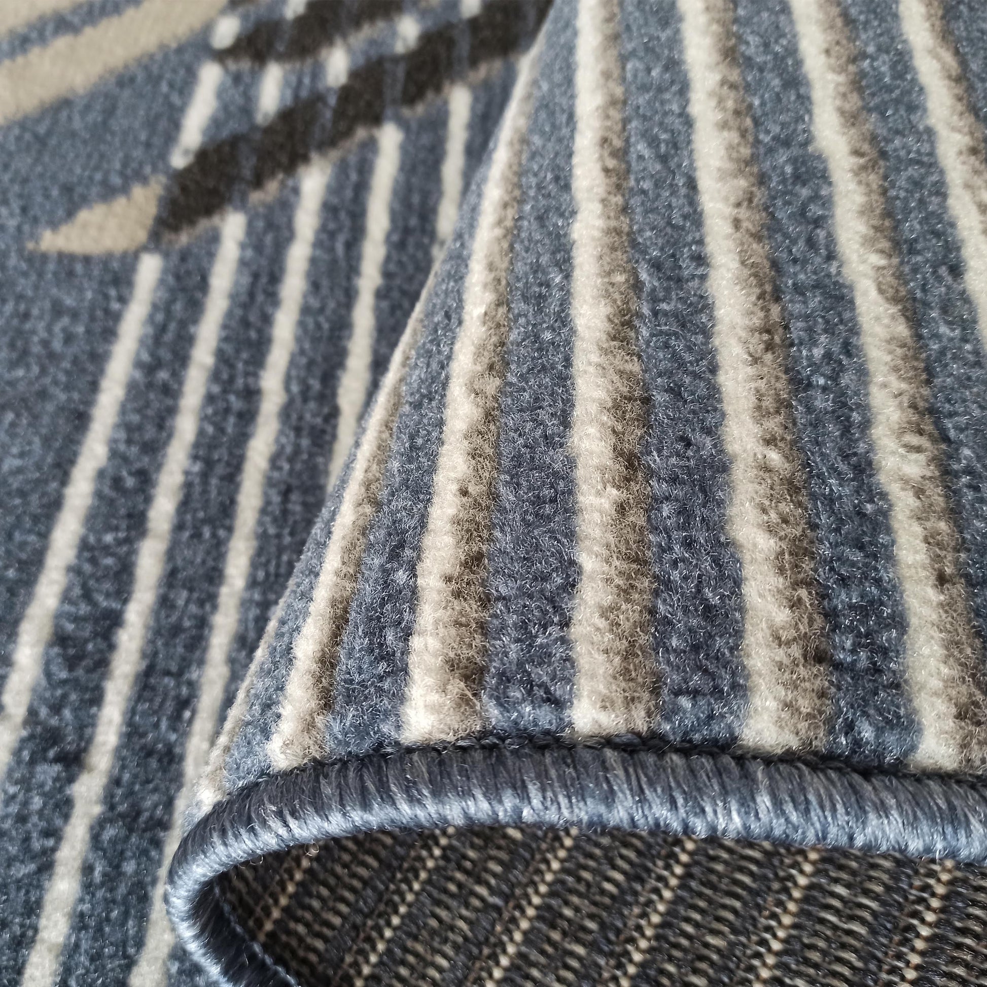 Tapis moelleux bleu gris, beige et blanc, avec des motifs géométriques et des rayures, l'un des côtés est roulé