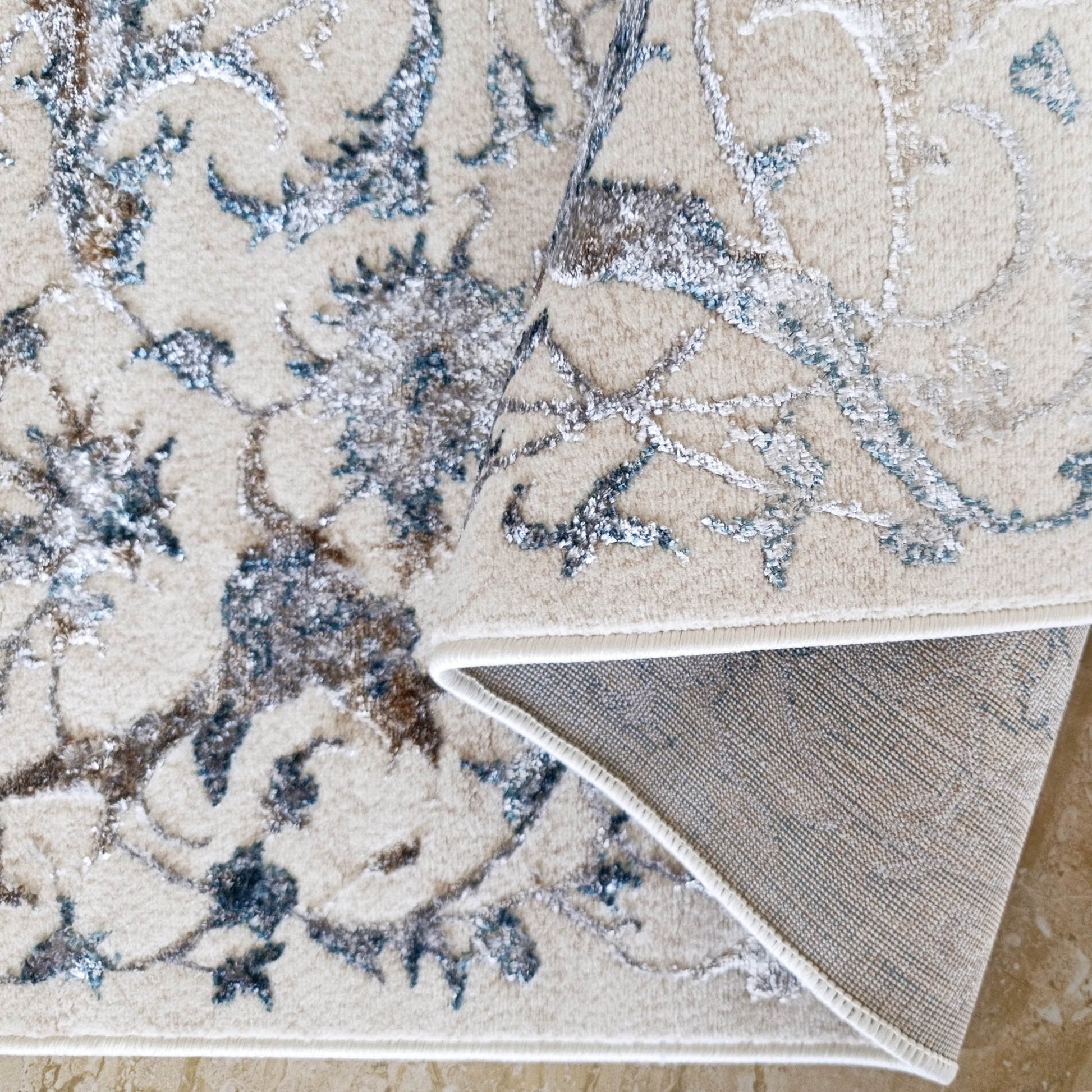 Tapis crème à motifs floraux bleus, gris et bruns, replié montrant l'envers tissé beige sur un sol clair