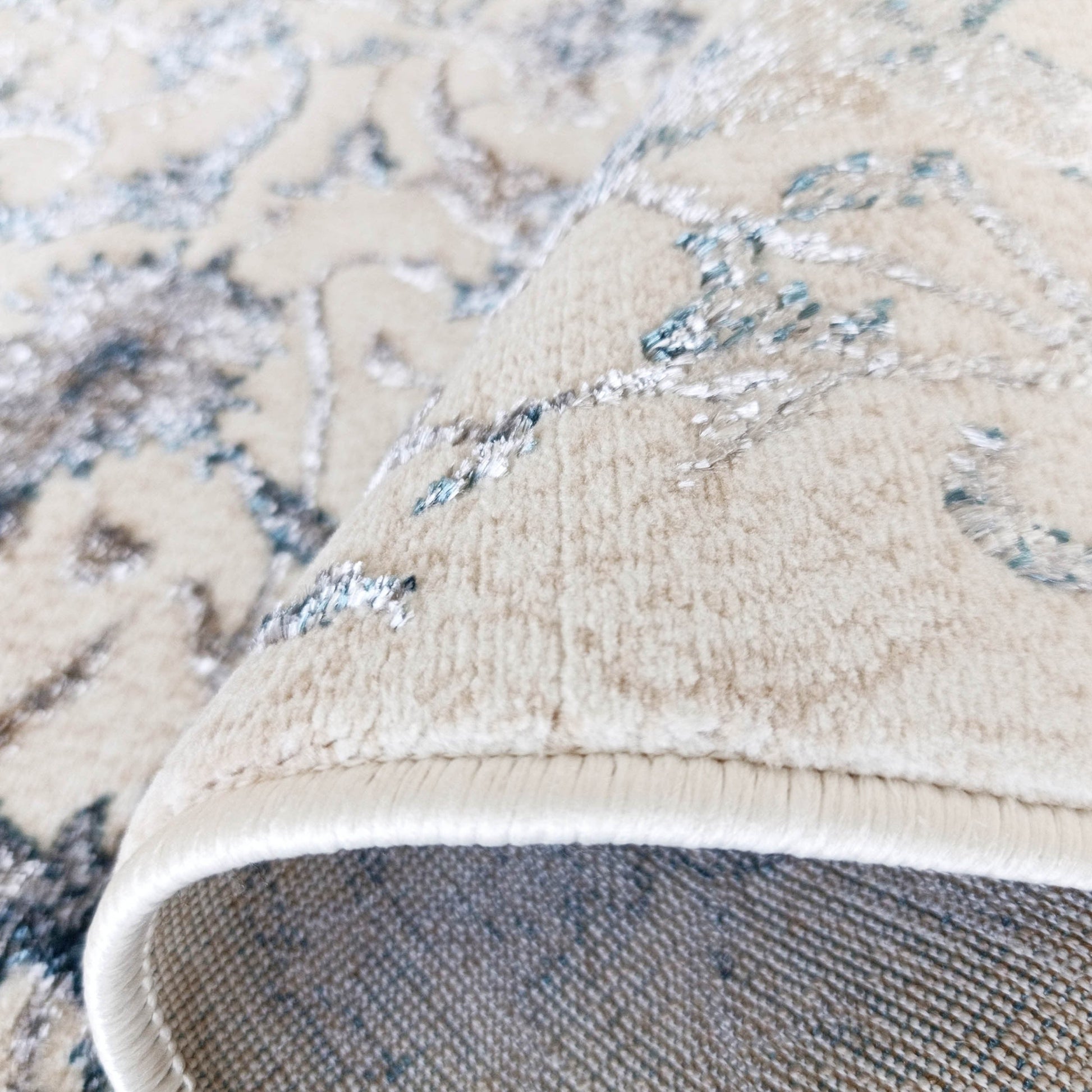 Tapis beige à motifs floraux bleus et gris, dont un coin est roulé, montrant la bordure blanche et le revers tissé gris