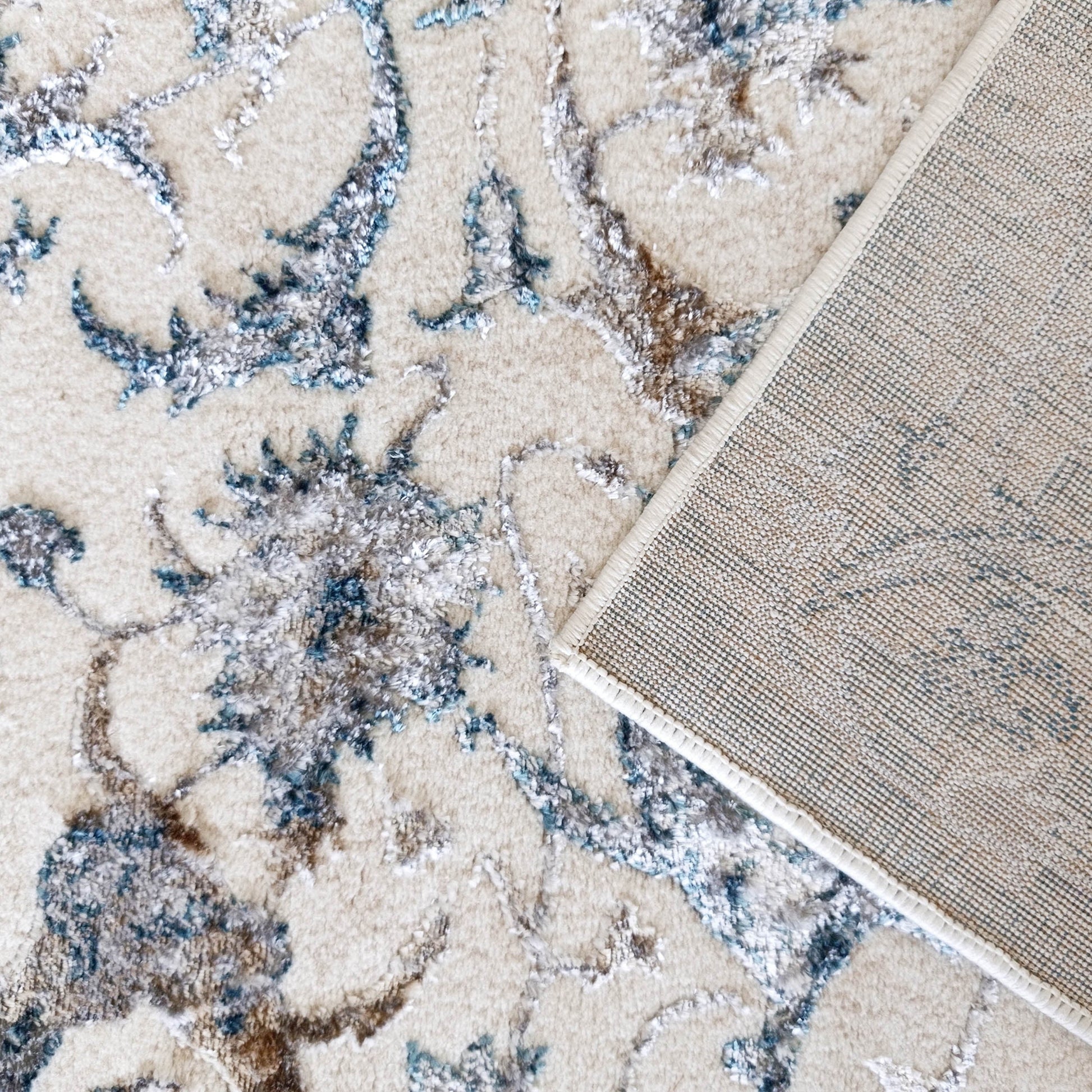 Tapis texturé écru avec des motifs floraux bleus, gris et bruns, et un coin replié révélant le tissage quadrillé de l'envers