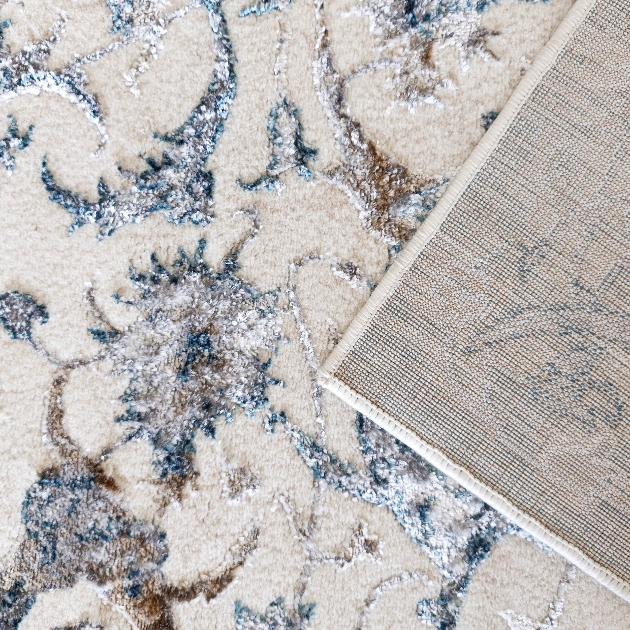 Tapis texturé écru avec des motifs floraux bleus, gris et bruns, et un coin replié révélant le tissage quadrillé de l'envers