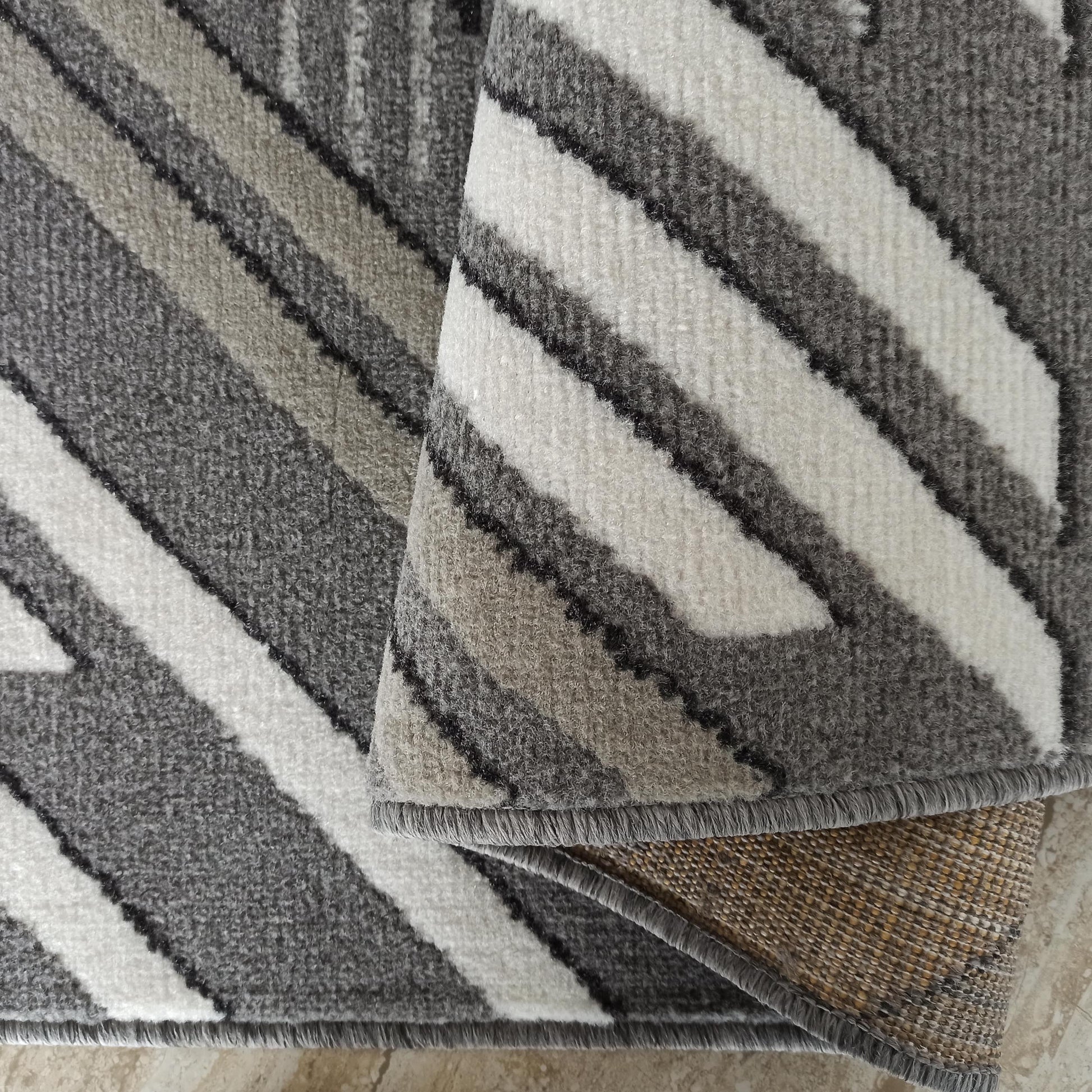 Tapis doux aux motifs géométriques rayés en gris, blanc et noir, avec une section enroulée qui révèle le revers tissé
