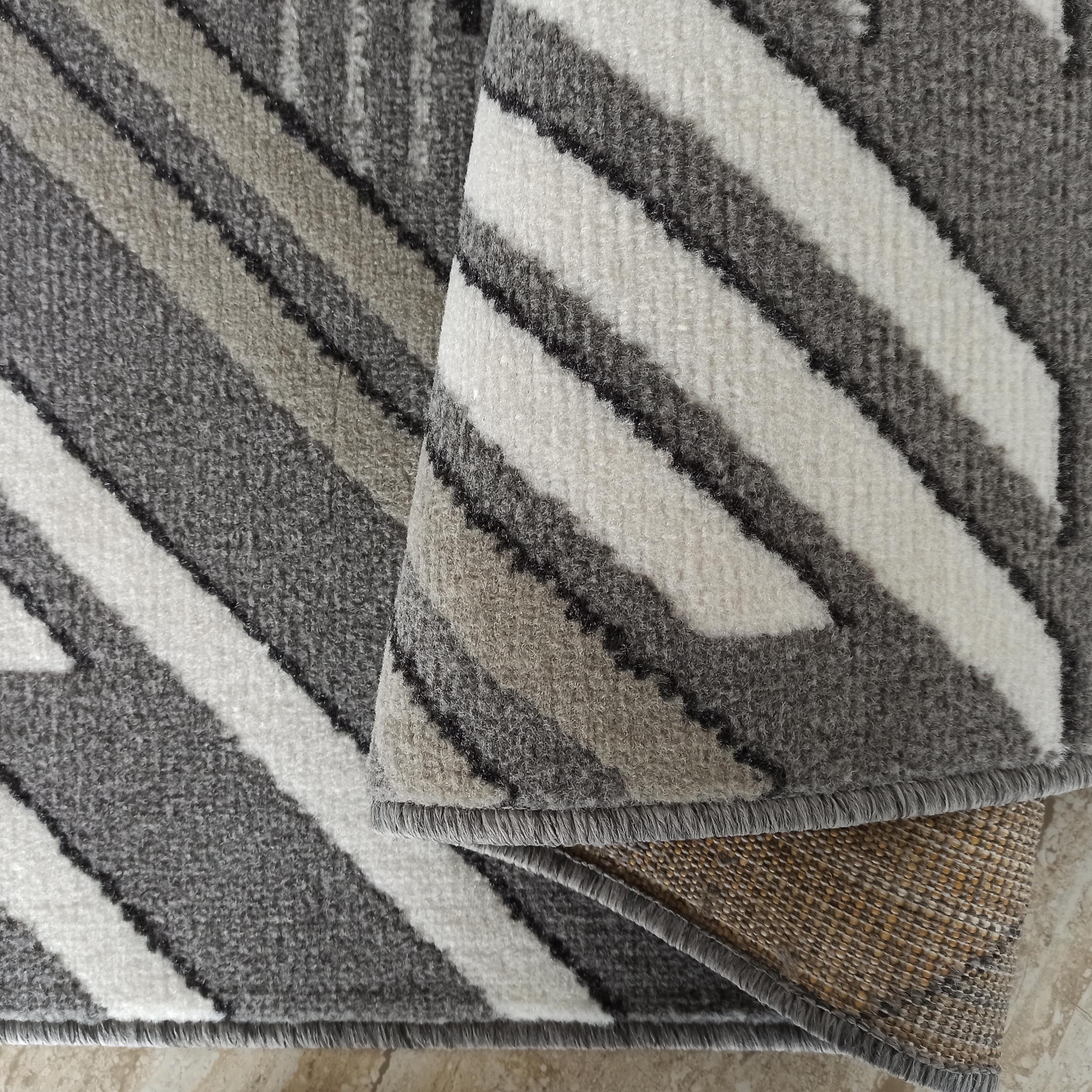 Tapis doux aux motifs géométriques rayés en gris, blanc et noir, avec une section enroulée qui révèle le revers tissé