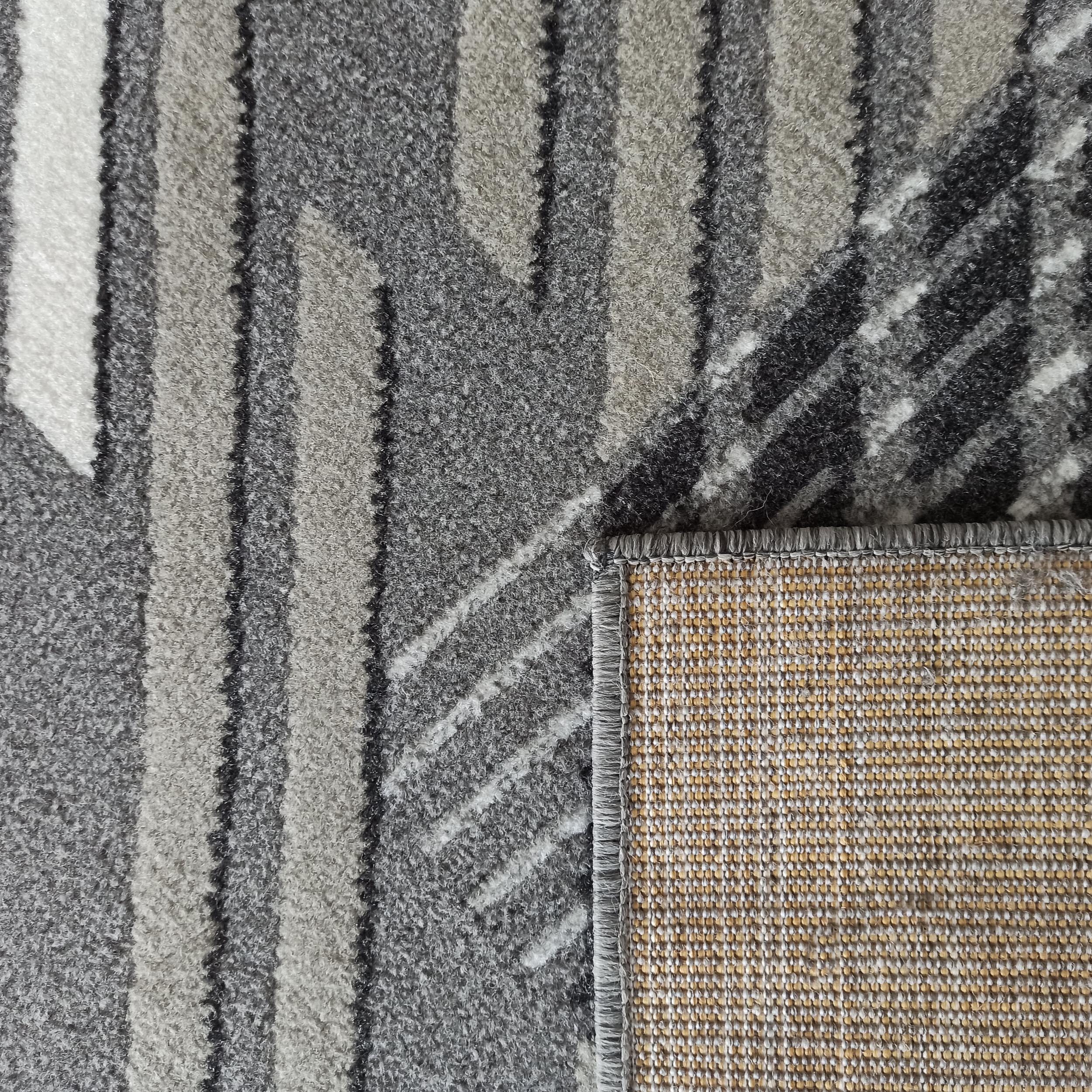 Tapis à poils doux aux motifs géométriques gris, noirs et blancs, montrant son envers en toile tissée beige et blanche