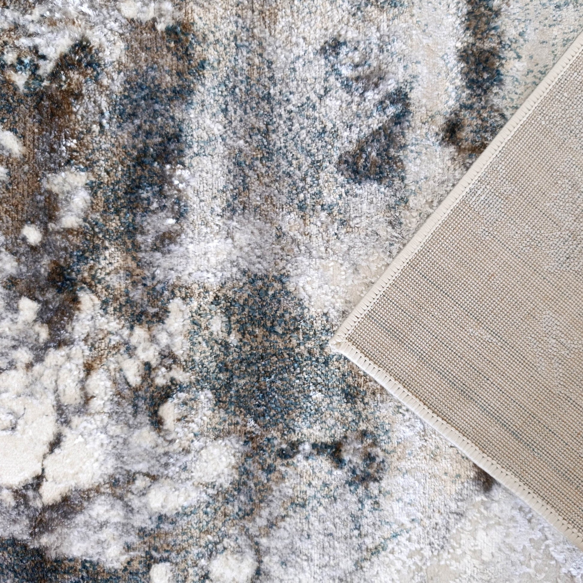 Tapis moelleux aux motifs abstraits crème, gris, marron et bleu-vert, dont un coin révèle le revers tissé beige clair