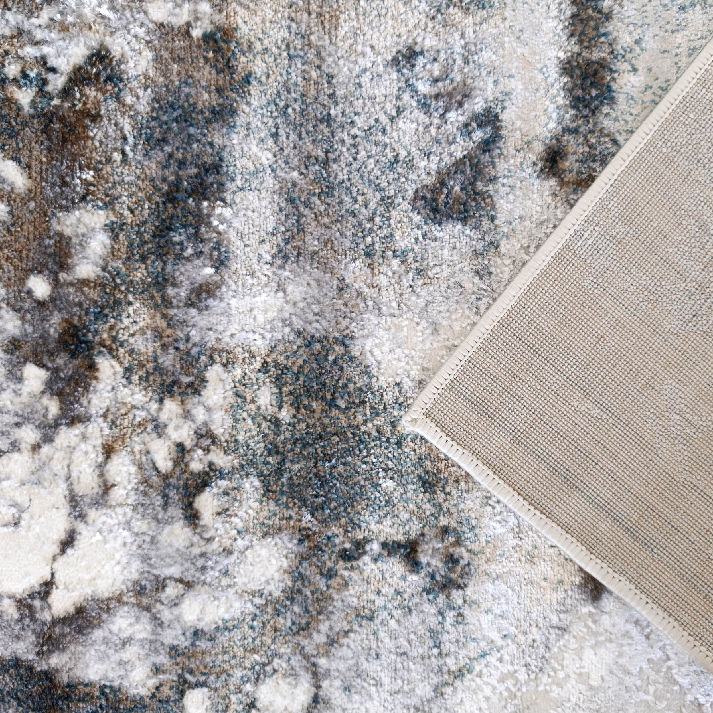 Tapis moelleux aux motifs abstraits crème, gris, marron et bleu-vert, dont un coin révèle le revers tissé beige clair