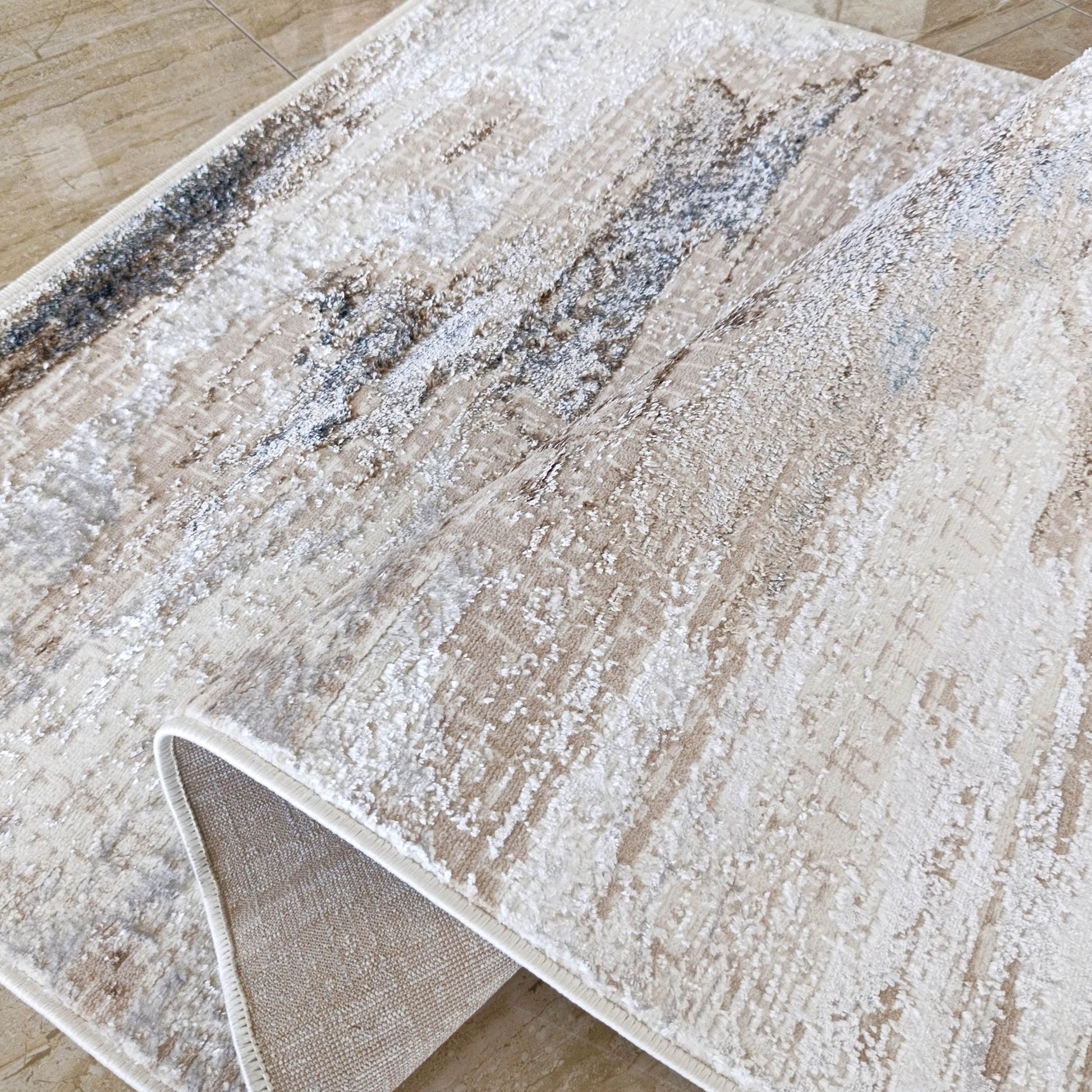 Tapis crème et beige aux touches abstraites gris foncé et bleu, roulé sur carrelage marron, dos tissé visible