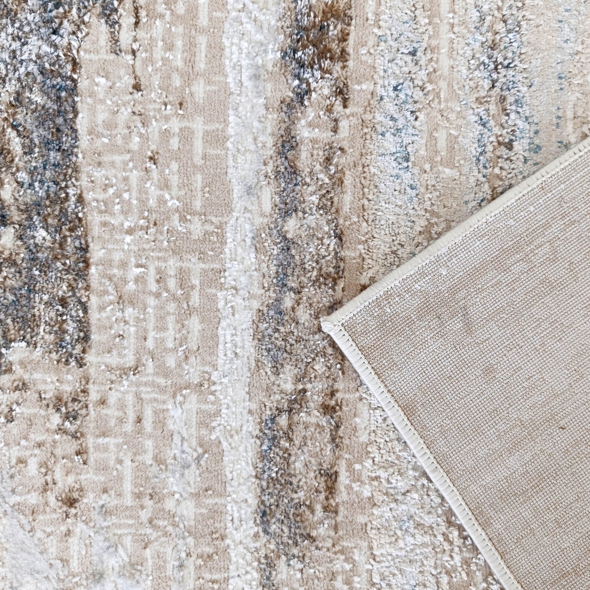 Tapis texturé aux motifs abstraits beige, marron et bleu. Un coin est relevé, révélant son envers en toile tissée claire