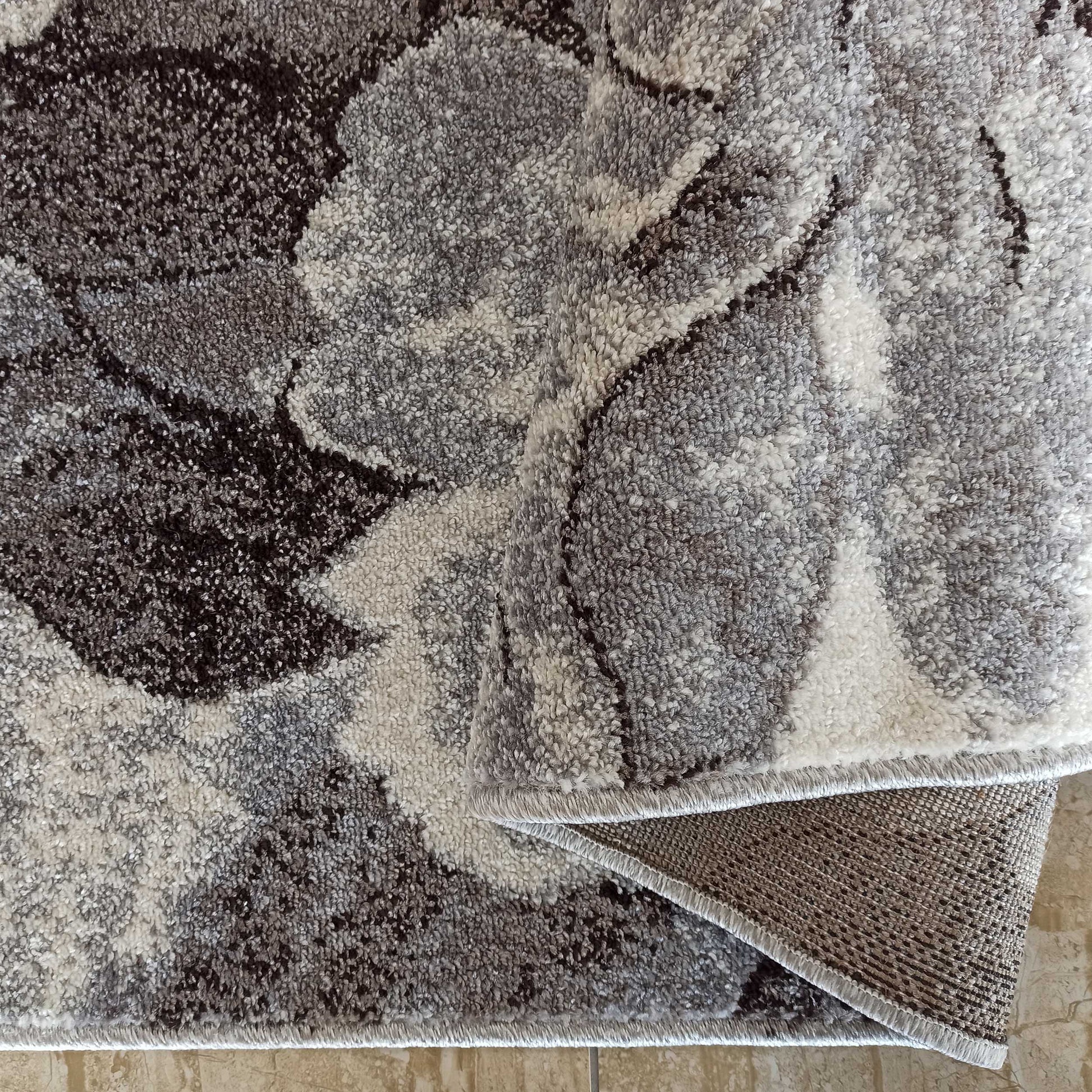 Tapis à poils longs gris, blancs et foncés avec des motifs floraux, partiellement roulé montrant son envers tissé sur un sol carrelé
