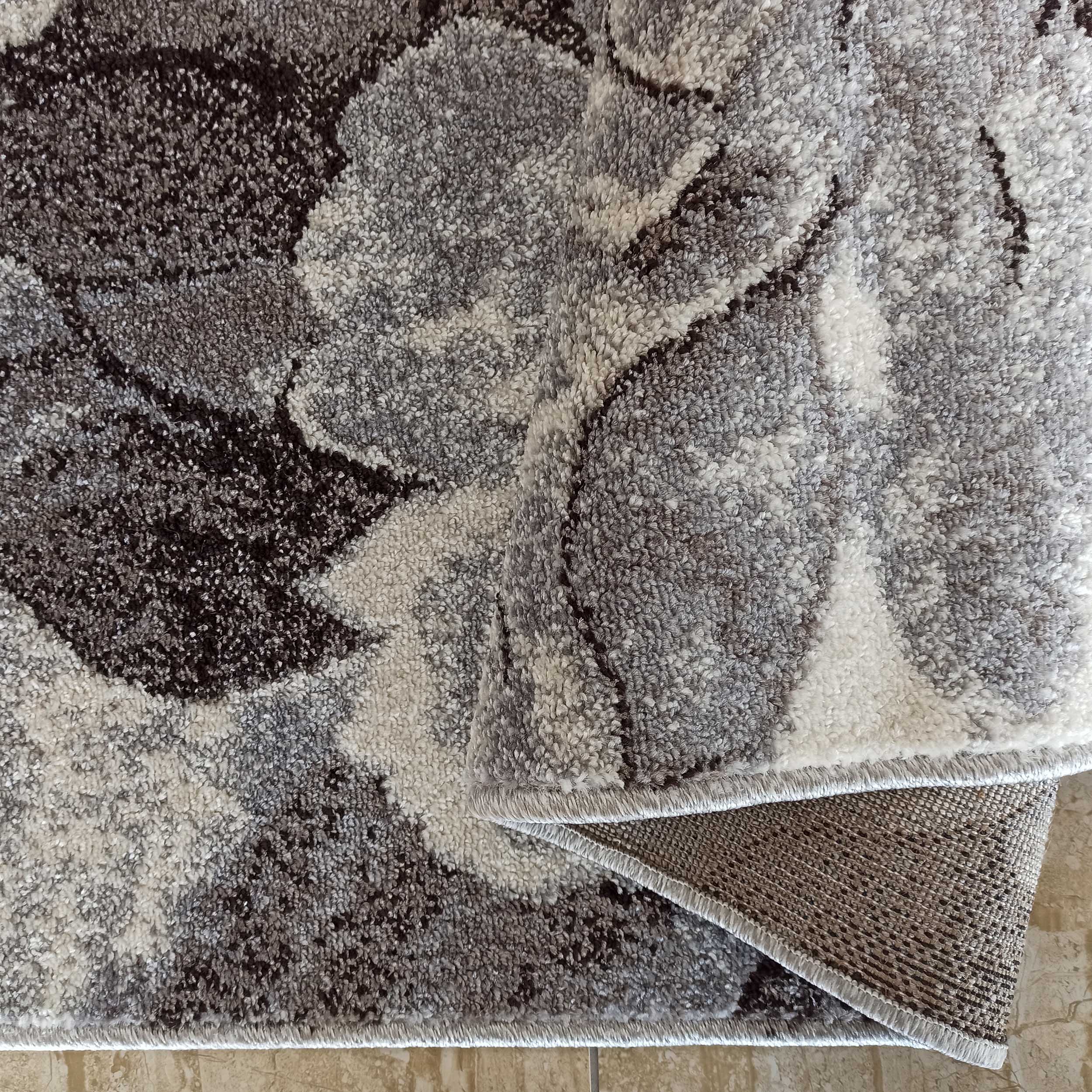 Tapis à poils longs gris, blancs et foncés avec des motifs floraux, partiellement roulé montrant son envers tissé sur un sol carrelé