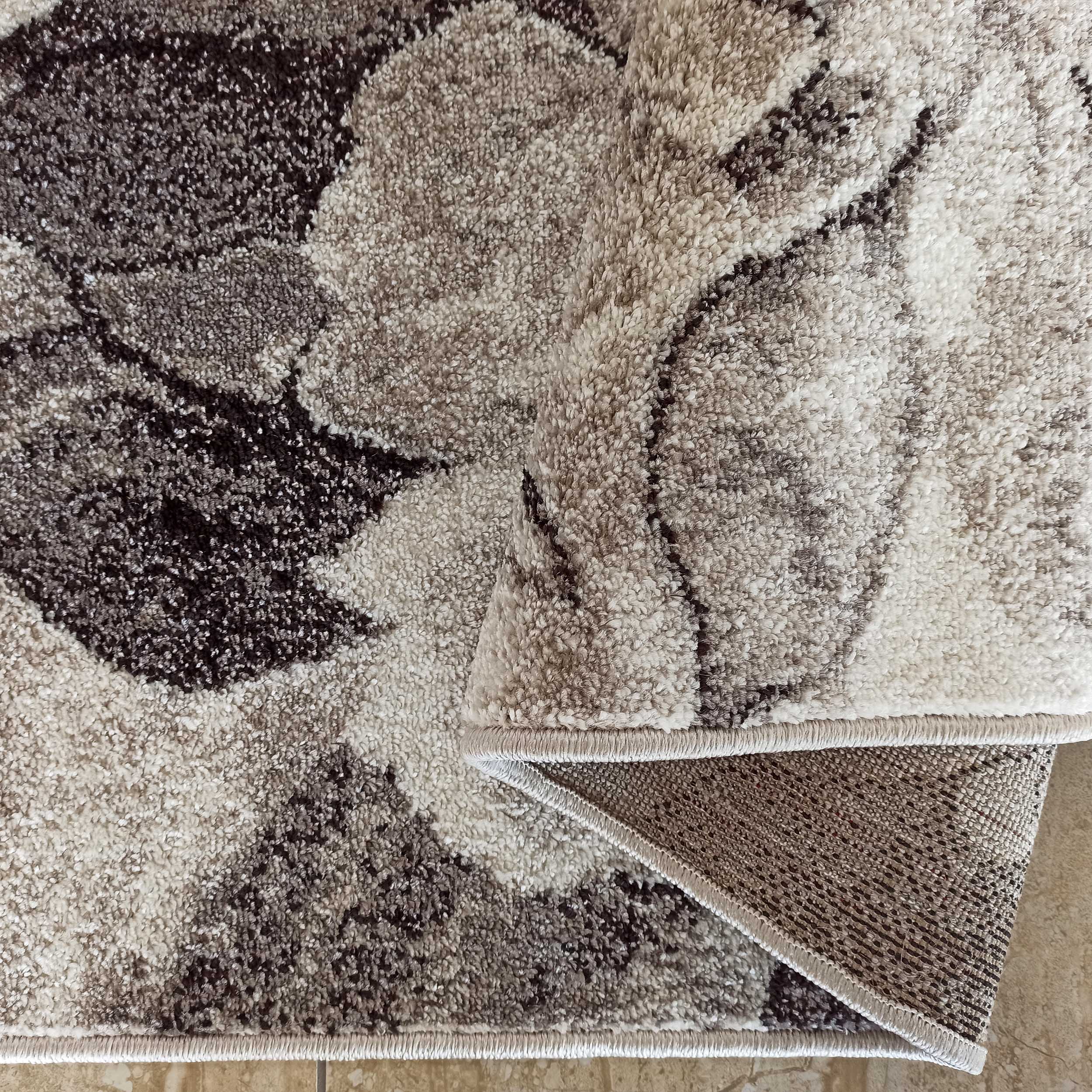 Tapis moelleux aux motifs tachetés abstraits en camaïeu de crème, beige et brun foncé, un coin est replié sur un sol carrelé