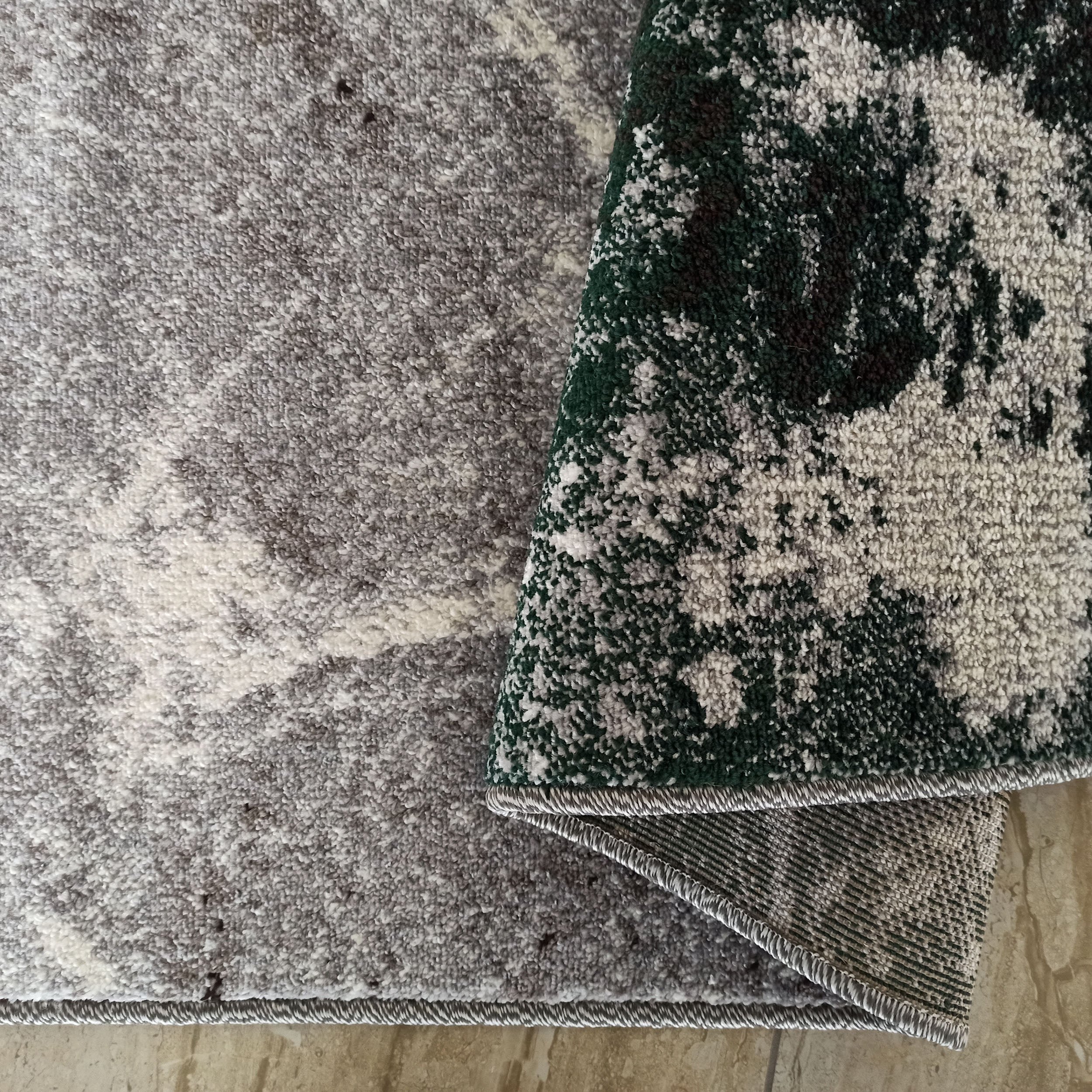 Tapis aux motifs abstraits gris, blanc et vert foncé. Une partie est repliée, laissant voir son envers tissé sur un carrelage