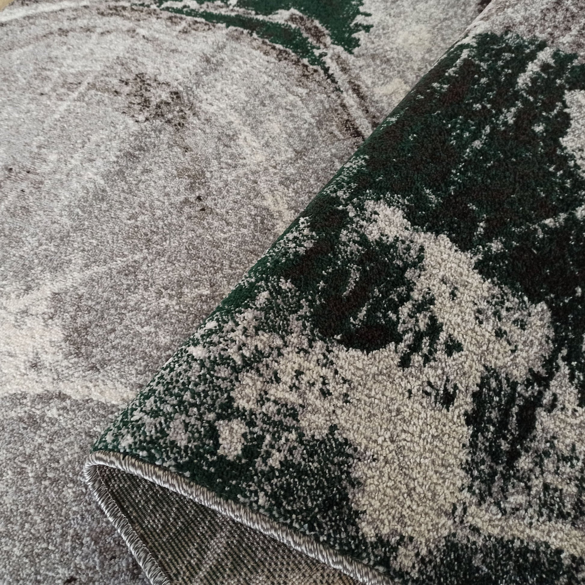 Tapis aux motifs abstraits mêlant gris clair, vert profond et touches de blanc, avec une surface texturée visible et une partie repliée