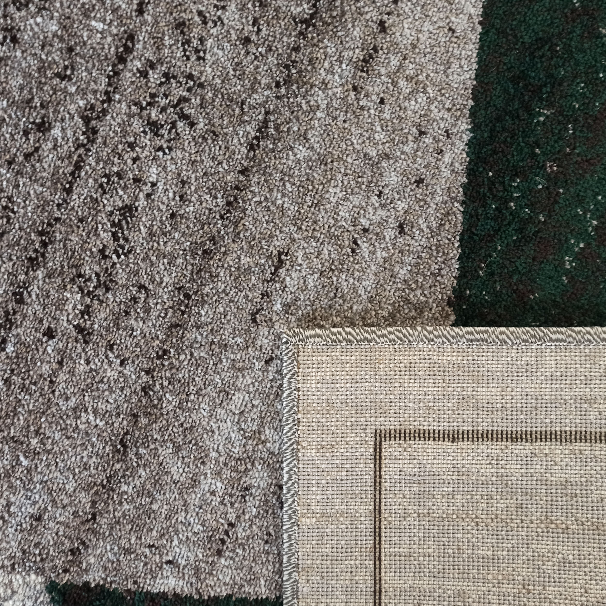 Un tapis gris et vert foncé à motifs mouchetés, avec un coin relevé montrant le dessous tissé beige et le bord surjeté