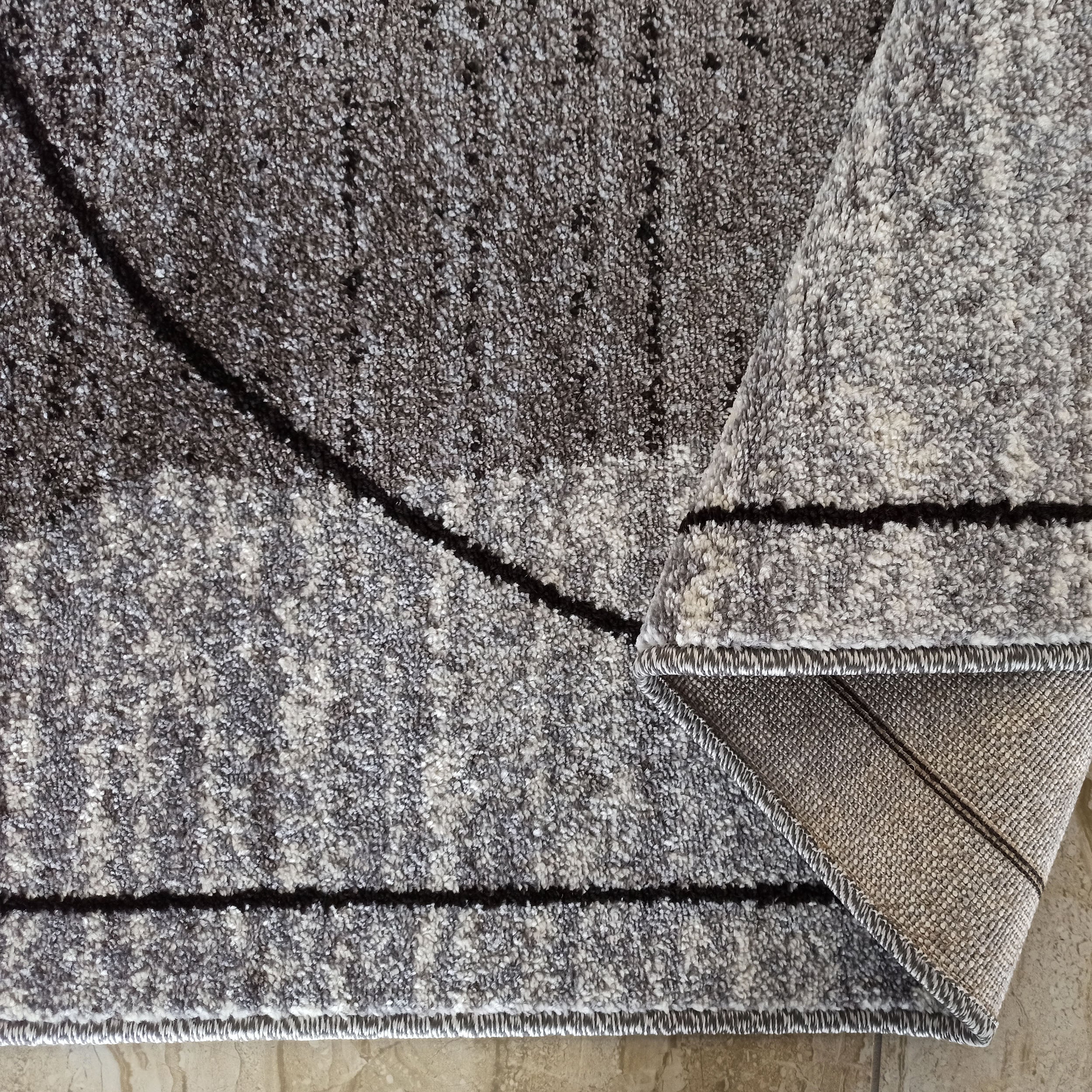 Tapis moelleux gris et blanc avec des motifs abstraits sombres, un coin est plié révélant l'envers tissé