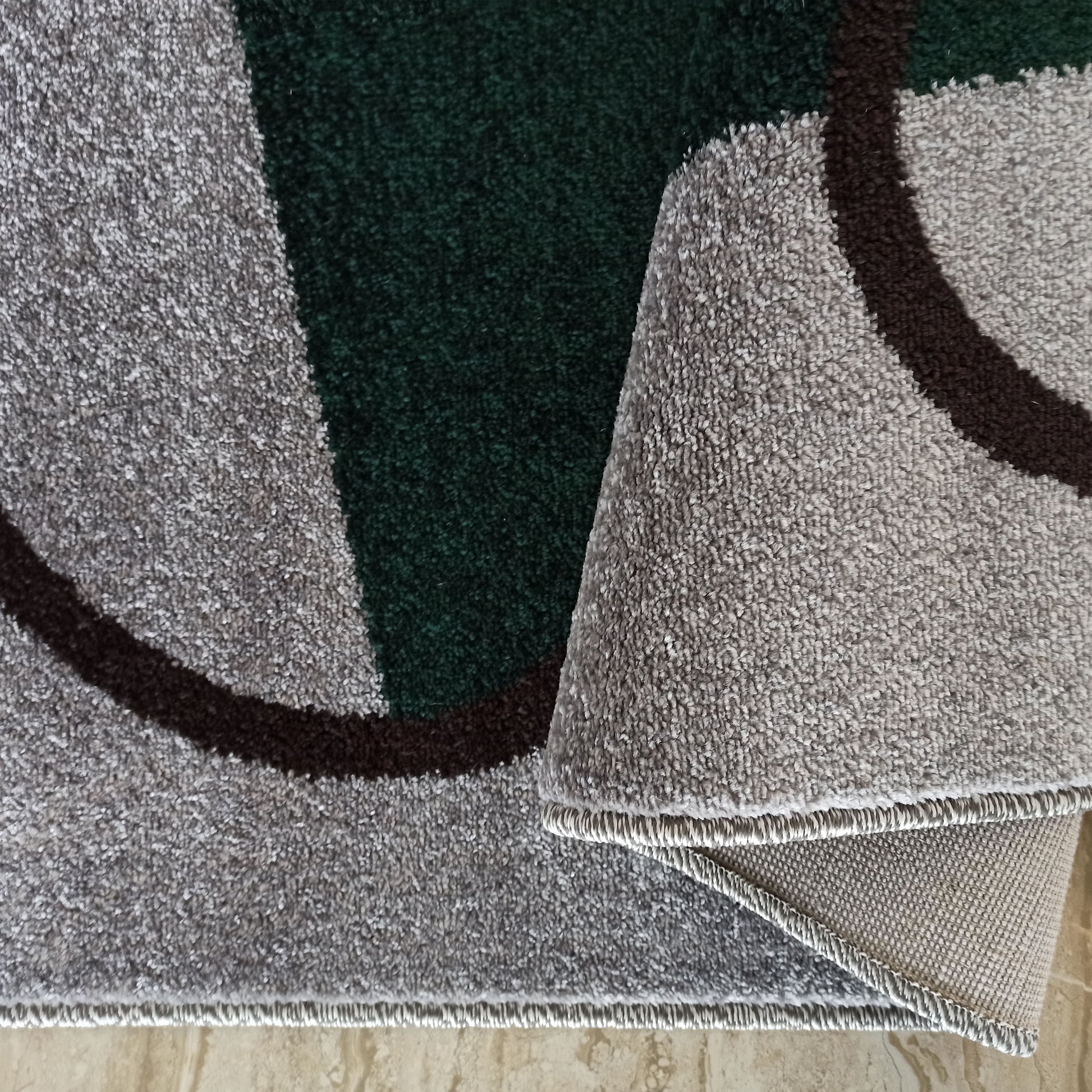 Tapis moelleux à motifs géométriques gris clair, vert forêt et marron foncé. Une section du tapis est pliée, révélant son envers