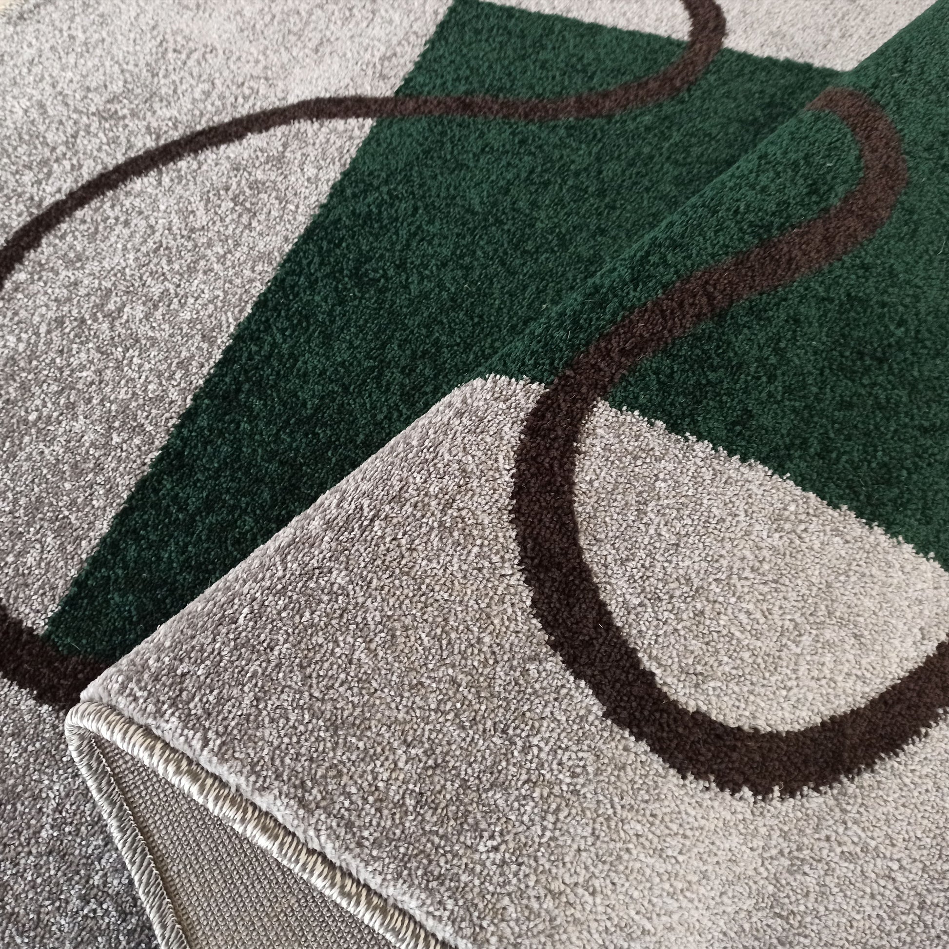 Tapis à motifs géométriques vert foncé et gris clair, avec des courbes marron. Sa texture est moelleuse et un bord est replié