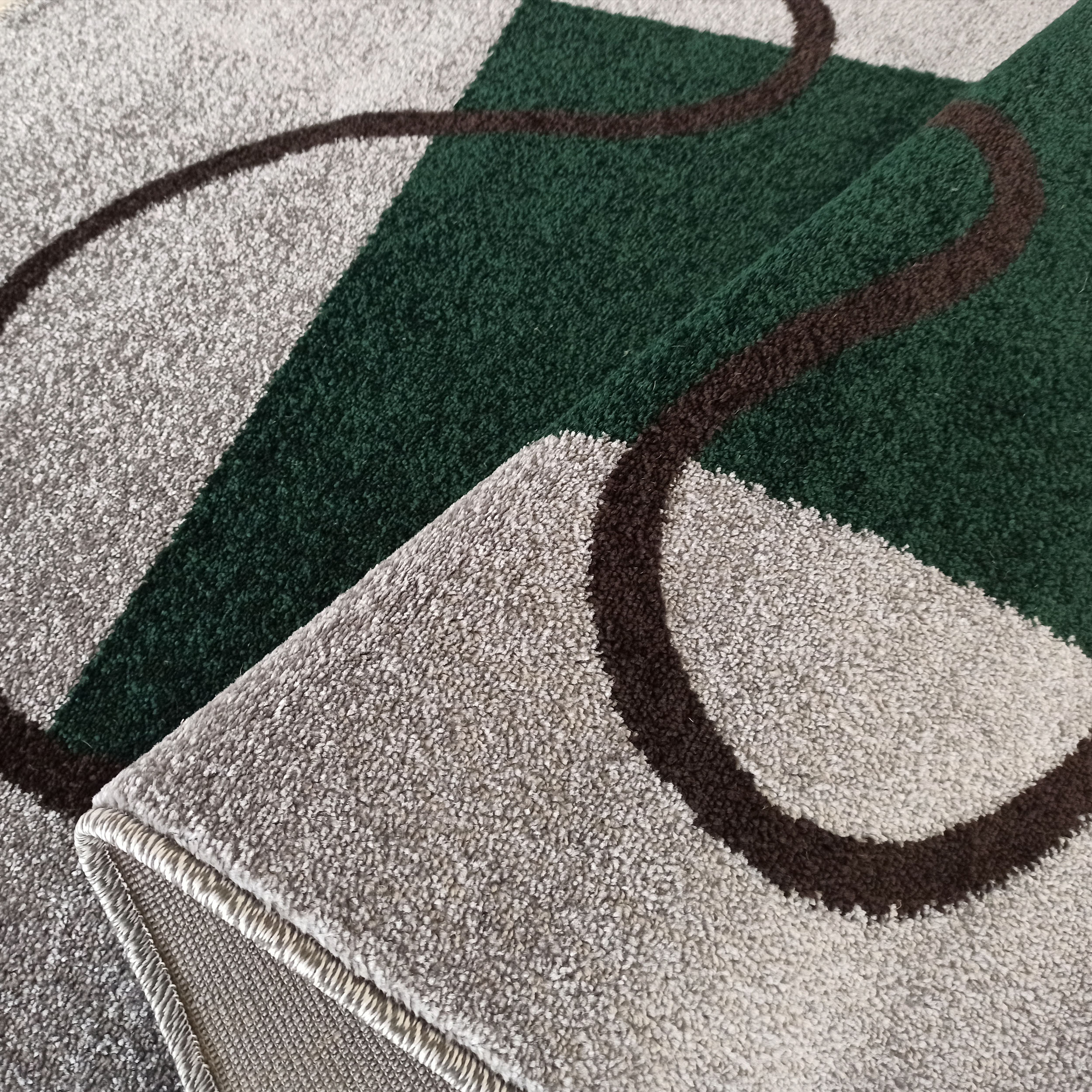 Tapis à motifs géométriques vert foncé et gris clair, avec des courbes marron. Sa texture est moelleuse et un bord est replié